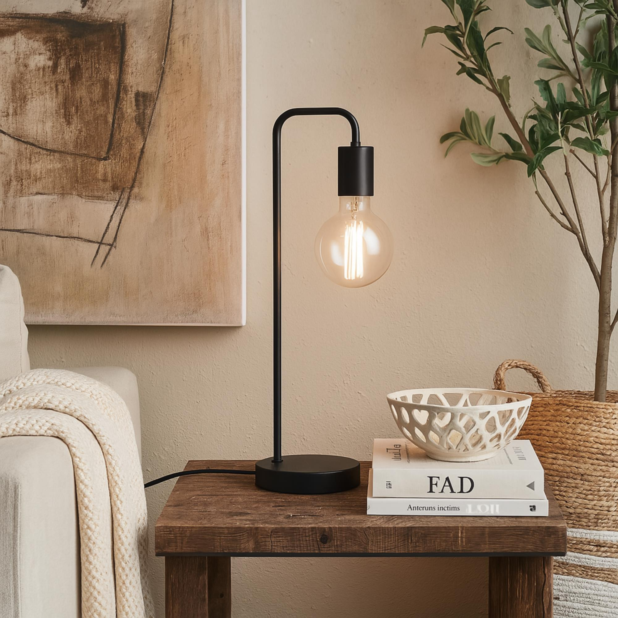 Una lámpara moderna con una bombilla grande se encuentra en una mesa de madera. Un cuenco blanco y libros están sobre la mesa. Un cuadro está al fondo.