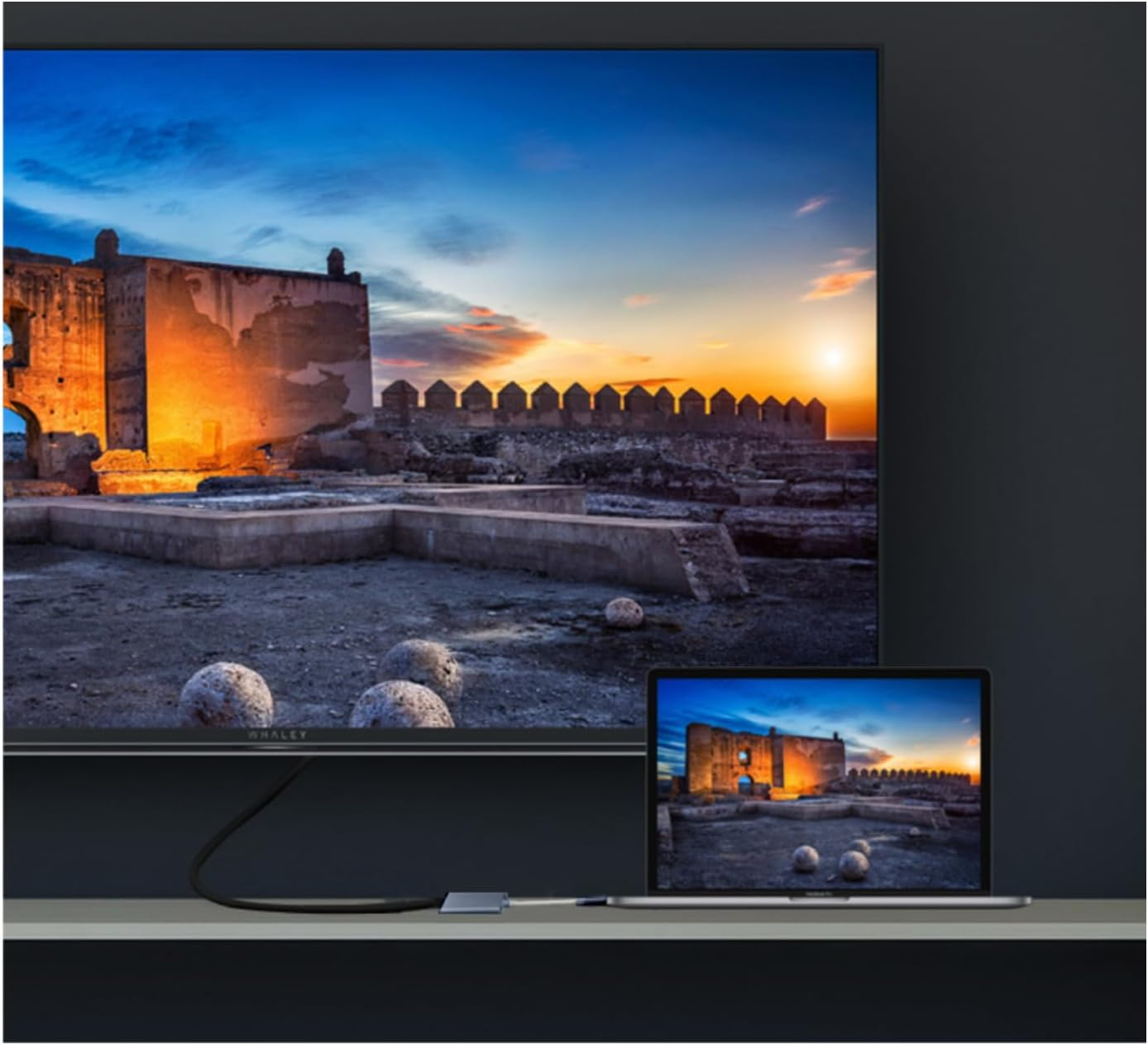 Un laptop e una TV mostrano la stessa scena del tramonto sopra antiche rovine. Un cavo li collega. Mensola grigia e sfondo scuro.
