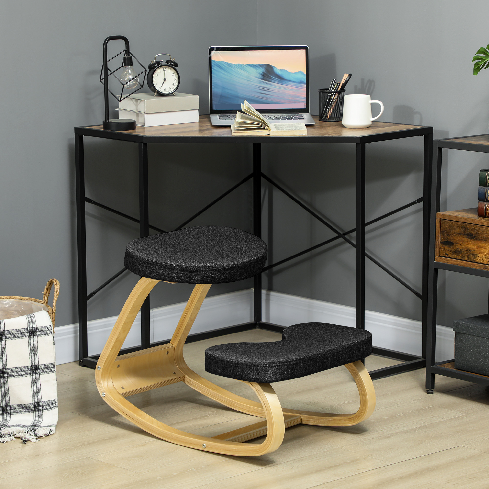 Una sedia inginocchiata è sotto una scrivania con un laptop e accessori. Sono visibili una lampada e una sveglia. Una coperta è in un cesto.