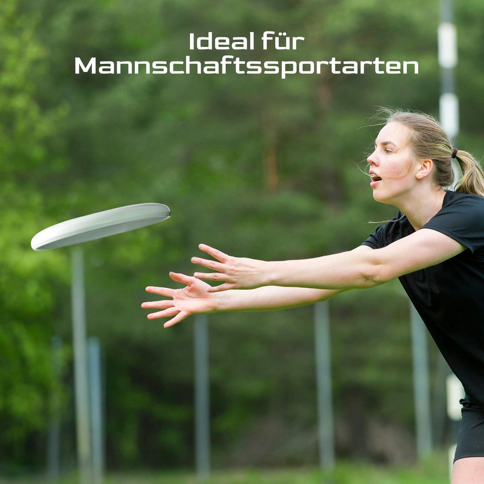 Eine Frau wirft eine Frisbee. Sie trägt schwarze Kleidung. Im Hintergrund sind grüne Bäume und ein Feld zu sehen.