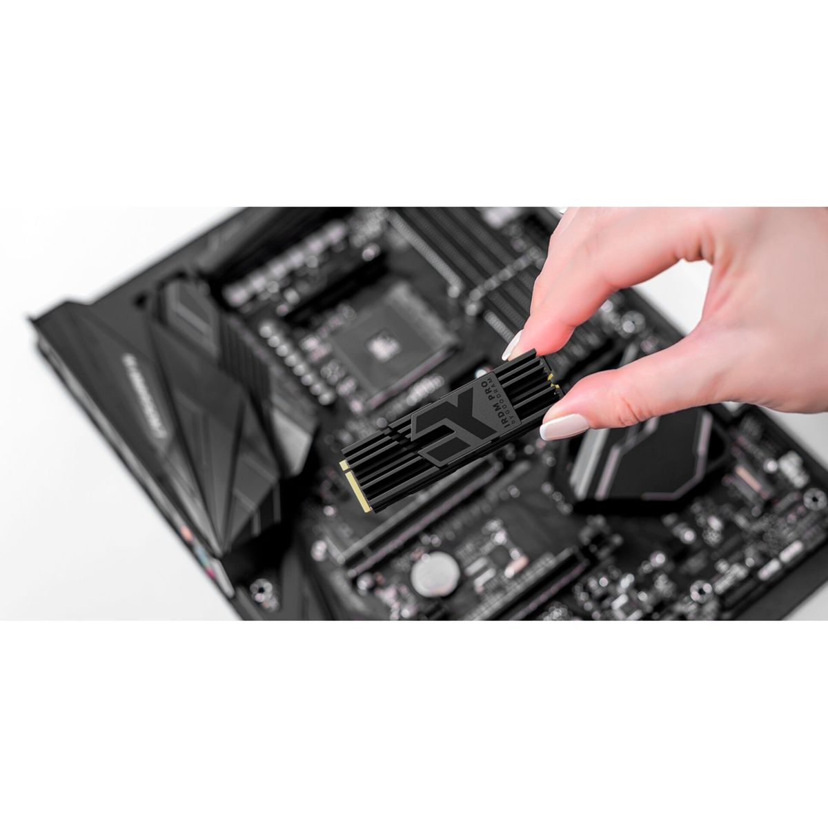 A hand holds a black SSD over a motherboard. The SSD has a metallic design. The motherboard is dark with various components.