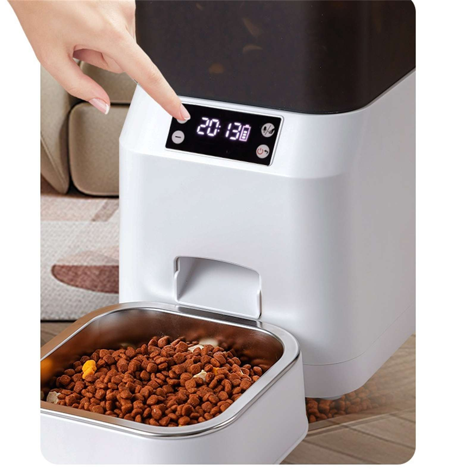 Eine Hand berührt einen weißen Haustierfutterautomaten mit digitaler Zeitanzeige. Eine Schüssel ist mit Tierfutter gefüllt.