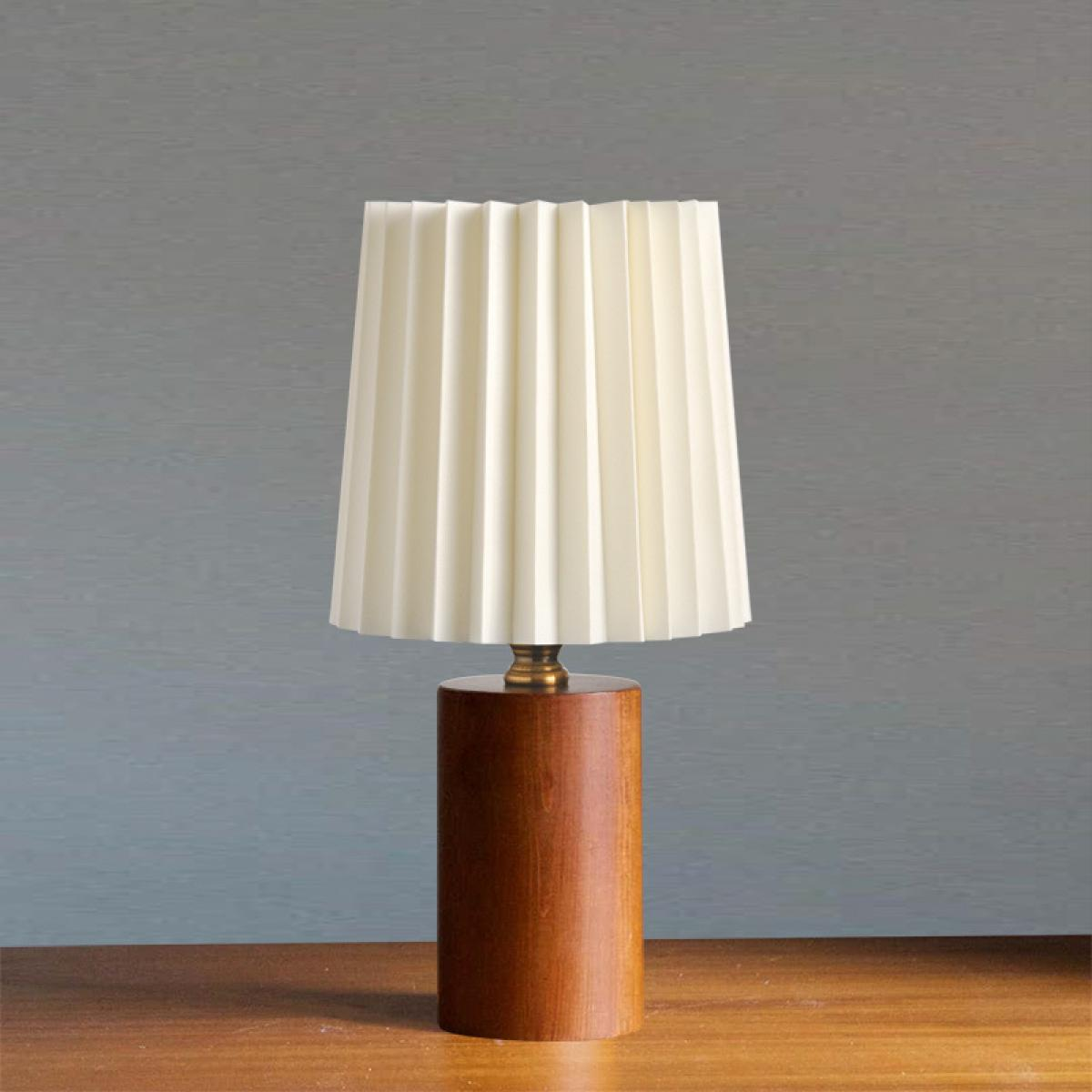 Eine Tischlampe mit cremefarbenem, plissiertem Lampenschirm steht auf einem Holzsockel. Der Hintergrund ist eine graue Wand.