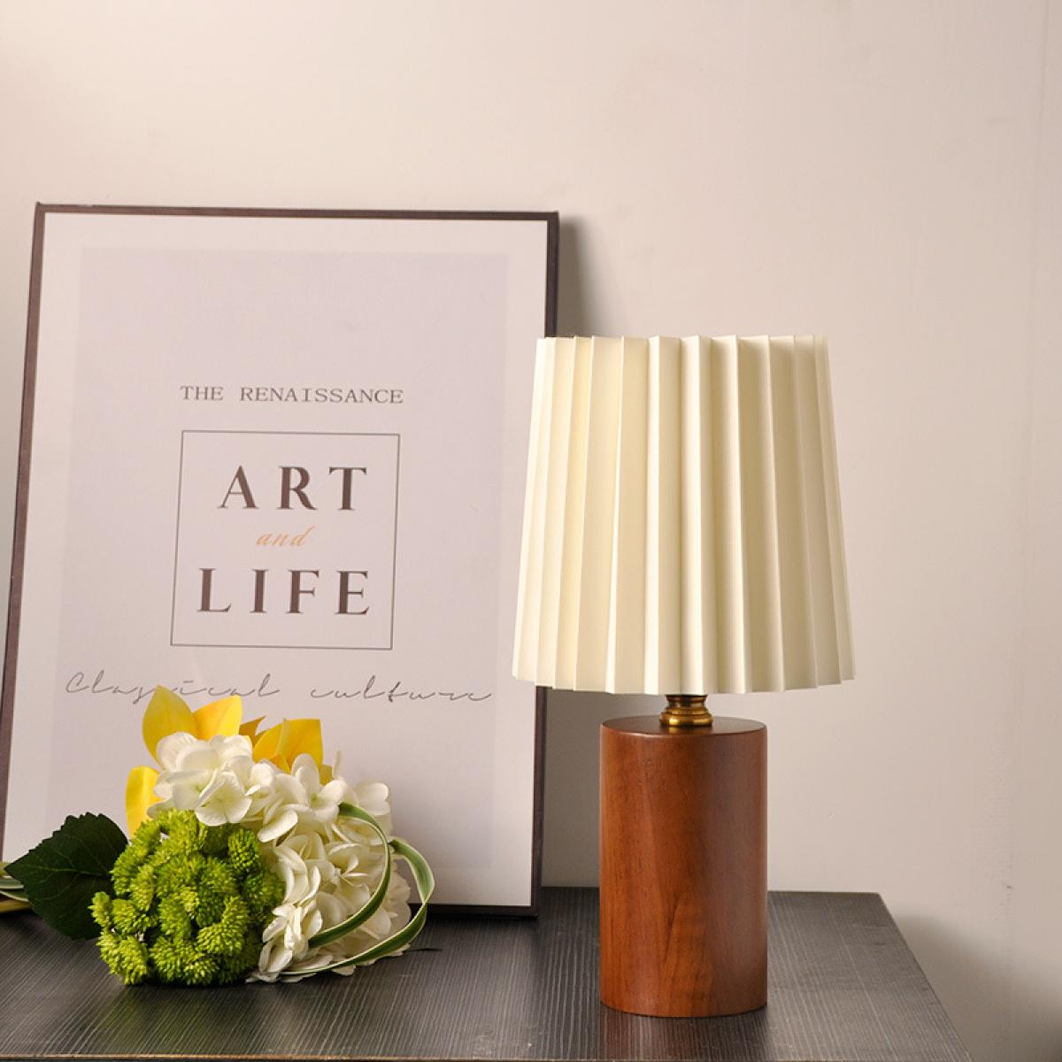 Eine Tischlampe mit plissiertem Lampenschirm steht auf einer dunklen Oberfläche. Ein gerahmter Kunstdruck und Blumen sind ebenfalls auf dem Tisch.