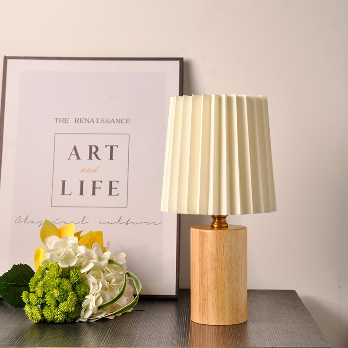 Eine Tischlampe mit cremefarbenem Schirm, Holzsockel und gerahmtem Kunstwerk. Blumen liegen neben der Lampe auf dem Tisch.
