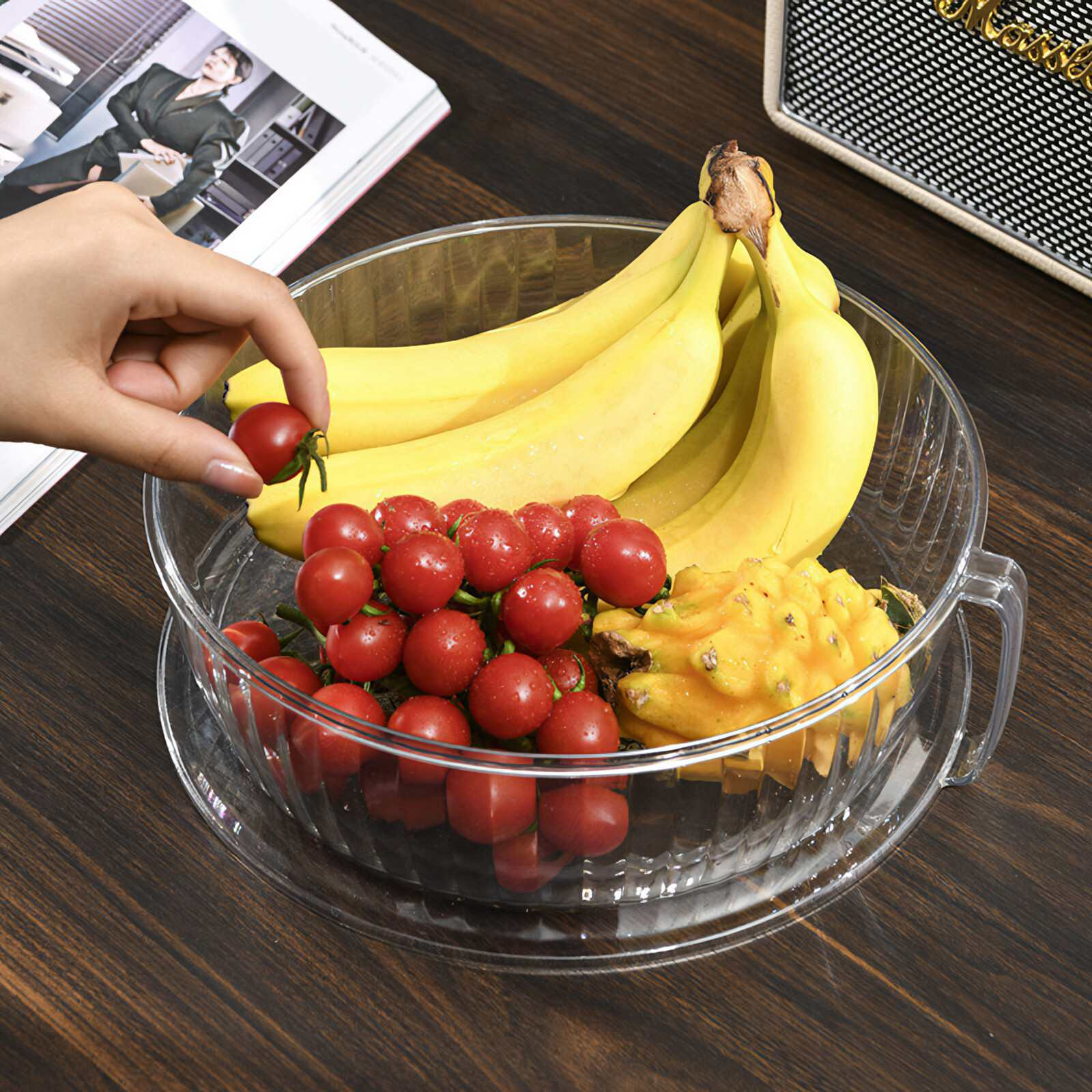Eine Hand hält eine Cherrytomate über einer Schüssel mit Früchten. Bananen, mehr Tomaten und eine gelbe Drachenfrucht sind ebenfalls in der Schüssel.