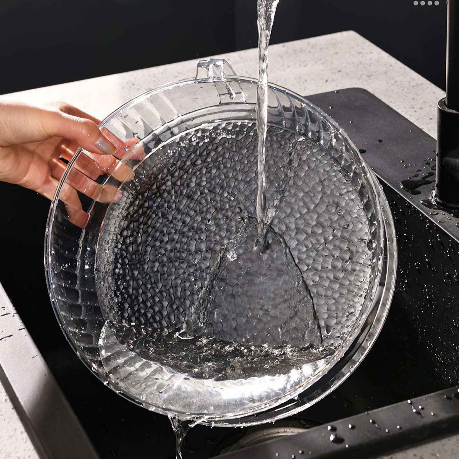 Wasser gießt in eine klare Schüssel mit strukturiertem Boden, die von einer Hand über einem schwarzen Waschbecken gehalten wird.
