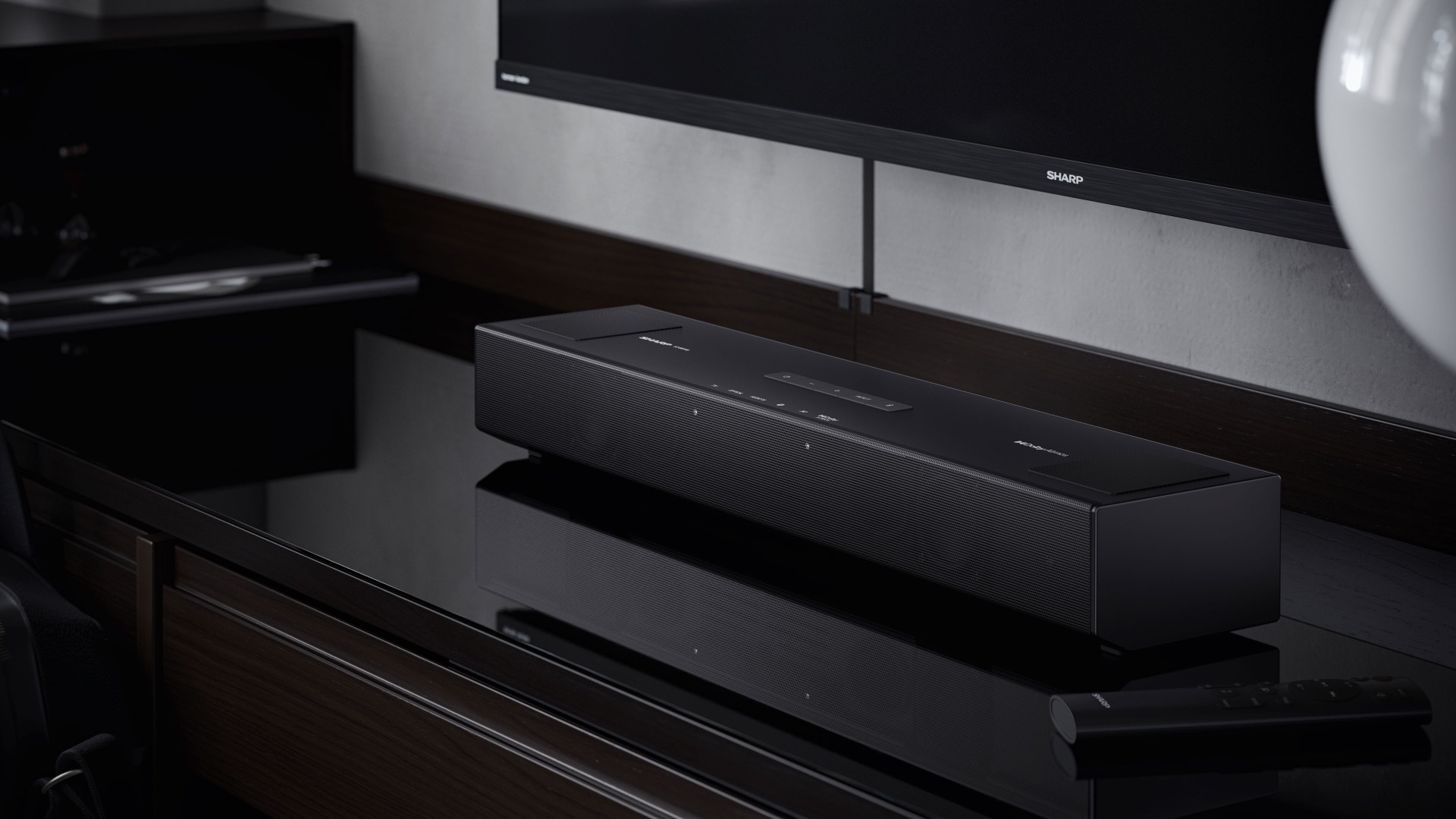 A black soundbar on a reflective surface. A TV and a black cabinet are in the background. A remote is visible.