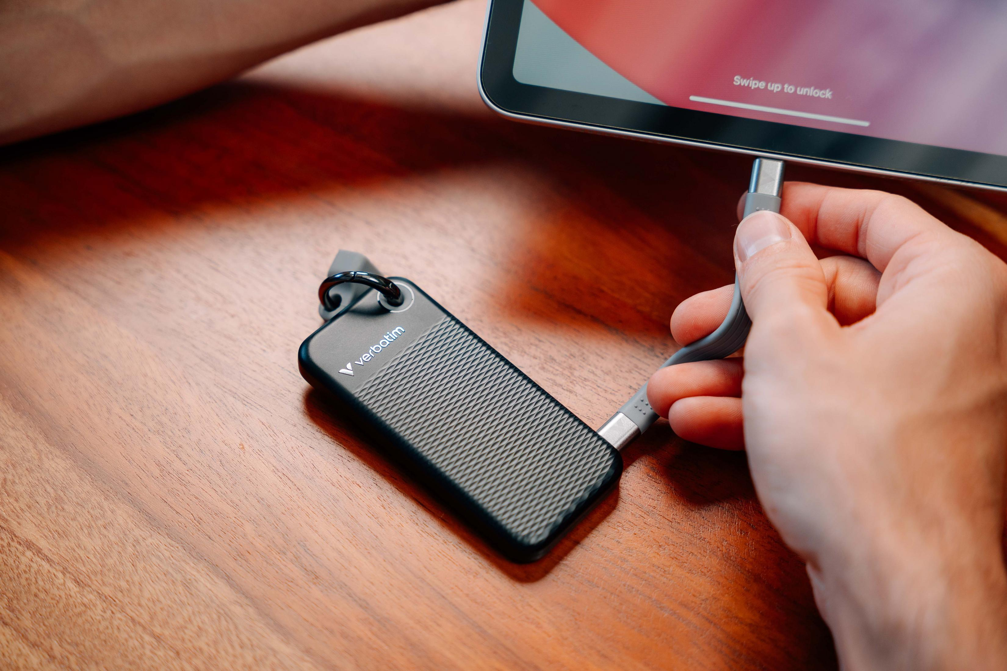 Ręka podłącza kabel USB-C do tabletu. Czarny dysk Verbatim z brelokiem leży na drewnianej powierzchni.