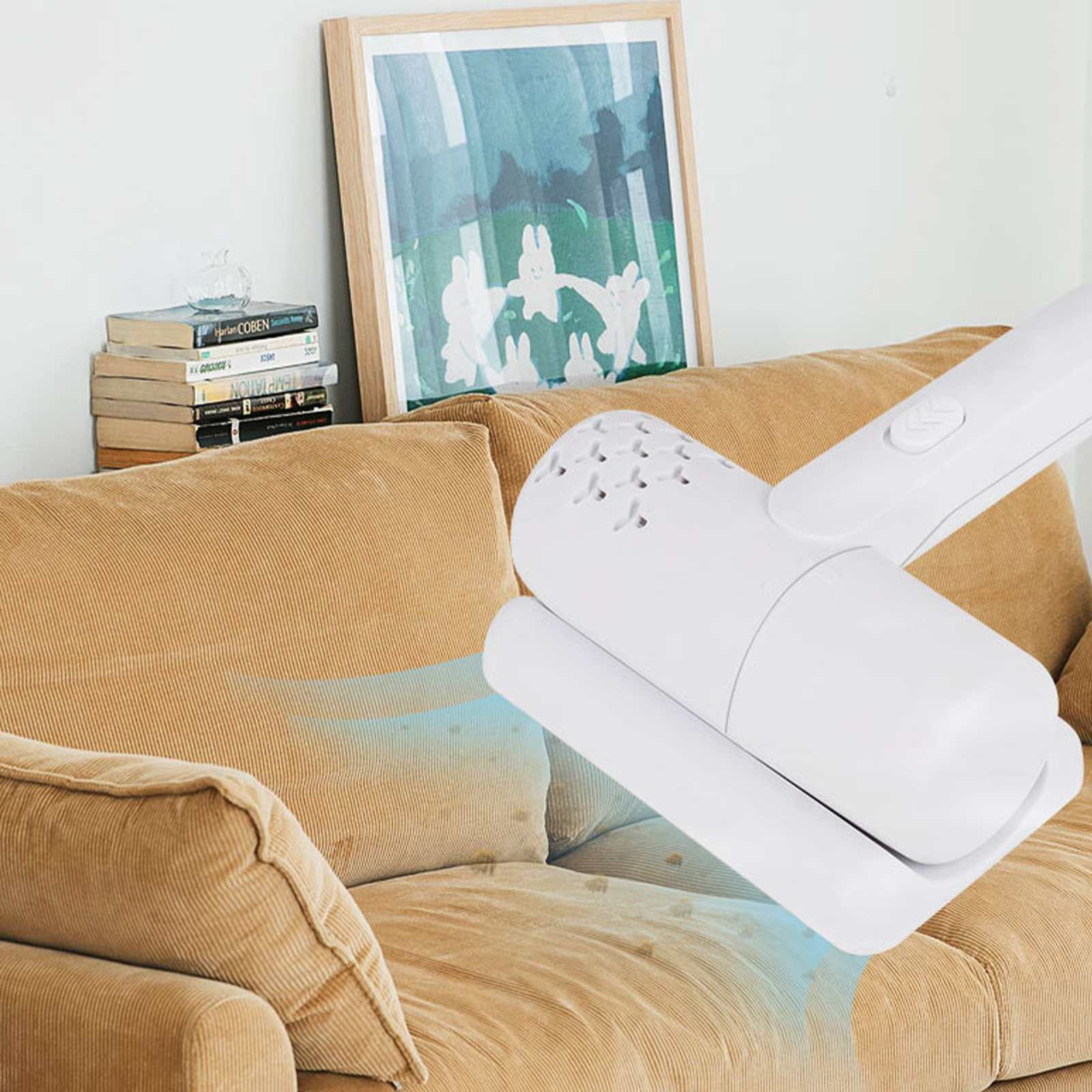 Ein weißer Staubsauger reinigt ein beiges Sofa. Im Hintergrund ein gerahmtes Bild und Bücher.