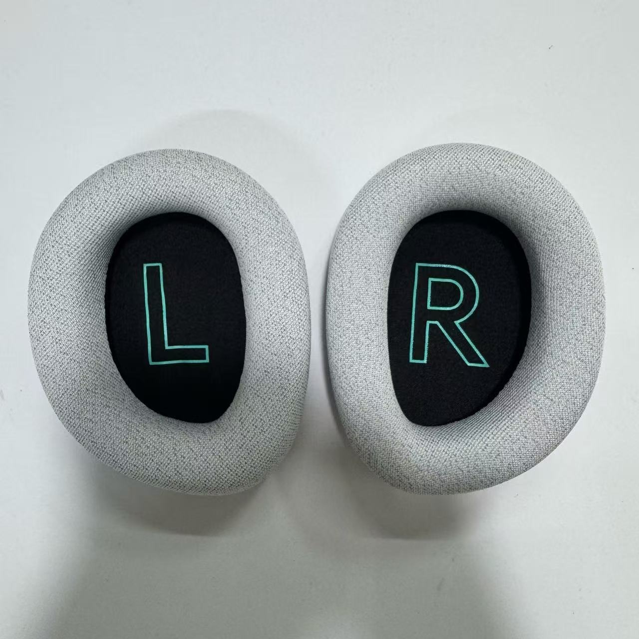 Zwei graue Ohrpolster, eines mit einem teal 'L', das andere mit einem teal 'R', auf weißem Untergrund.