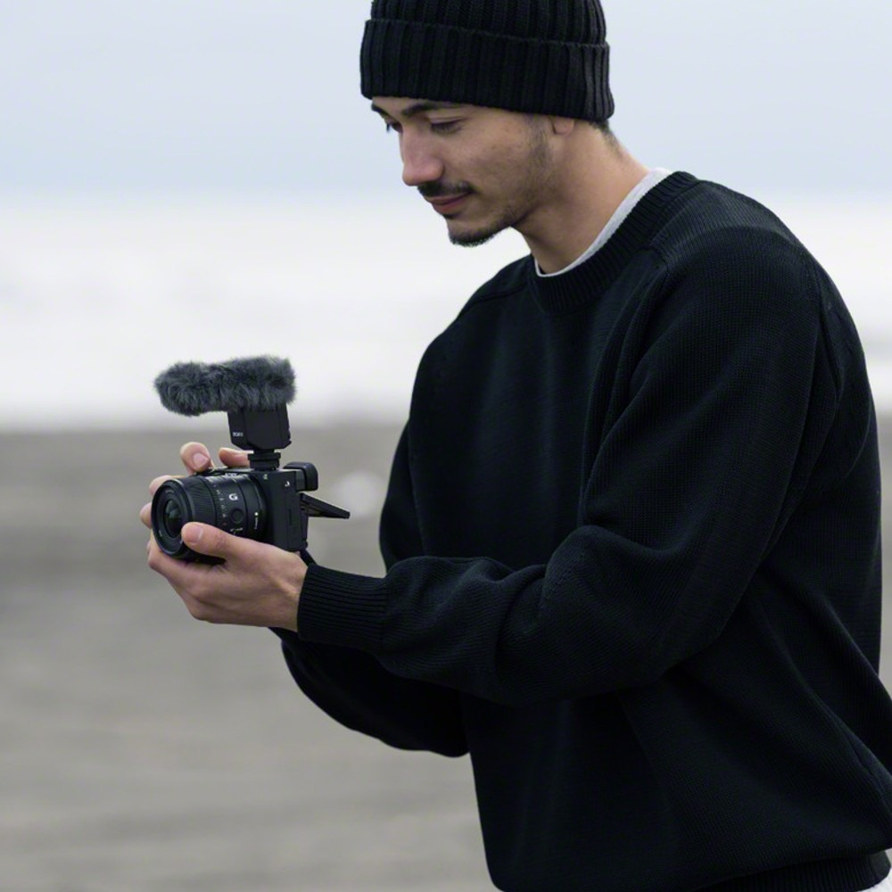 Mężczyzna w czarnym swetrze i czapce trzyma kamerę z mikrofonem. Patrzy na kamerę na plaży.