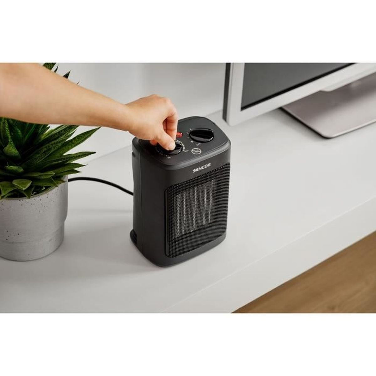 Una mano ajusta el dial de un calefactor Sencor negro. Una planta en maceta a la izquierda, un monitor a la derecha, todo sobre una superficie blanca.