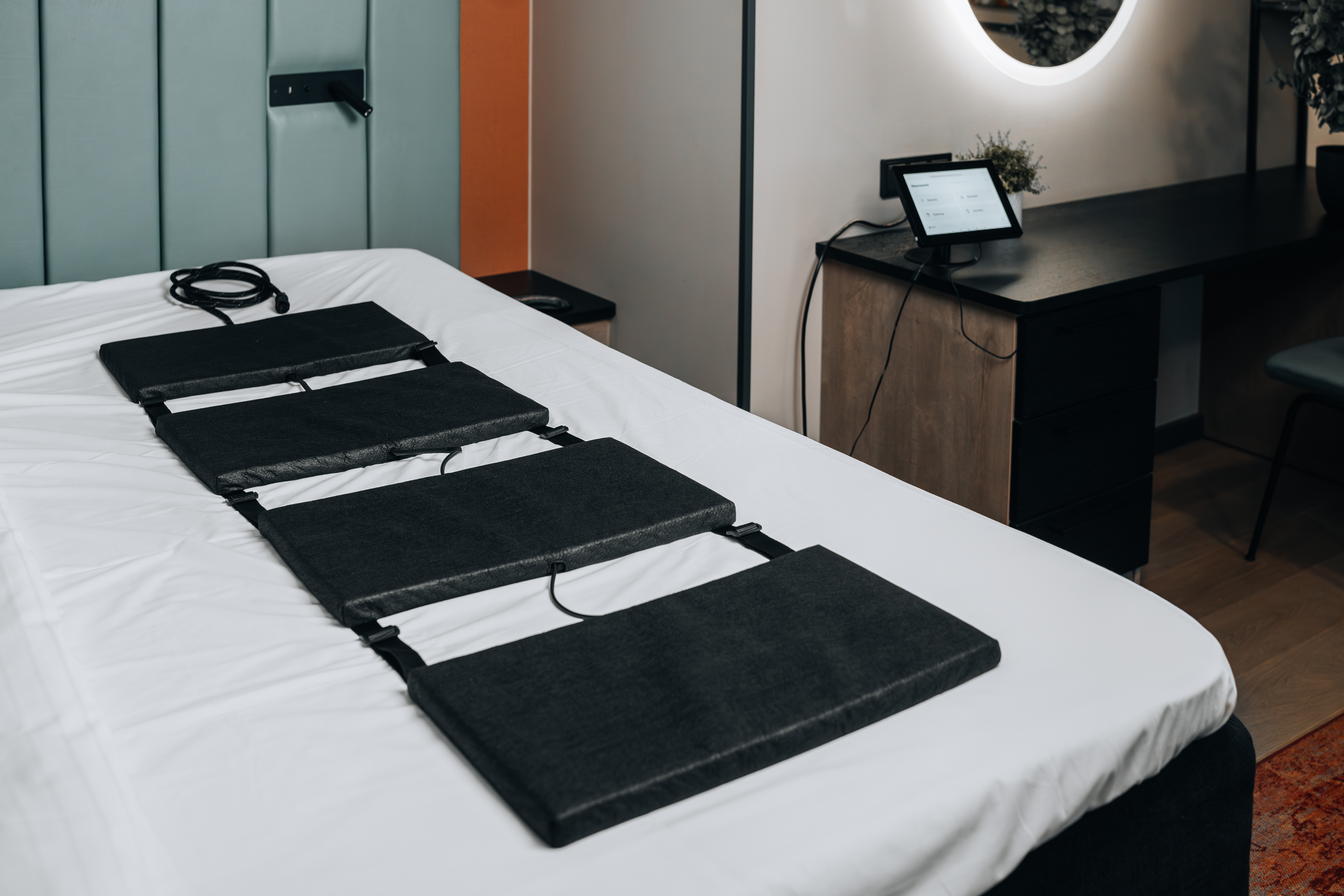 Een bed met een zwart massagekussen, een bureau met een tablet en een moderne lamp.