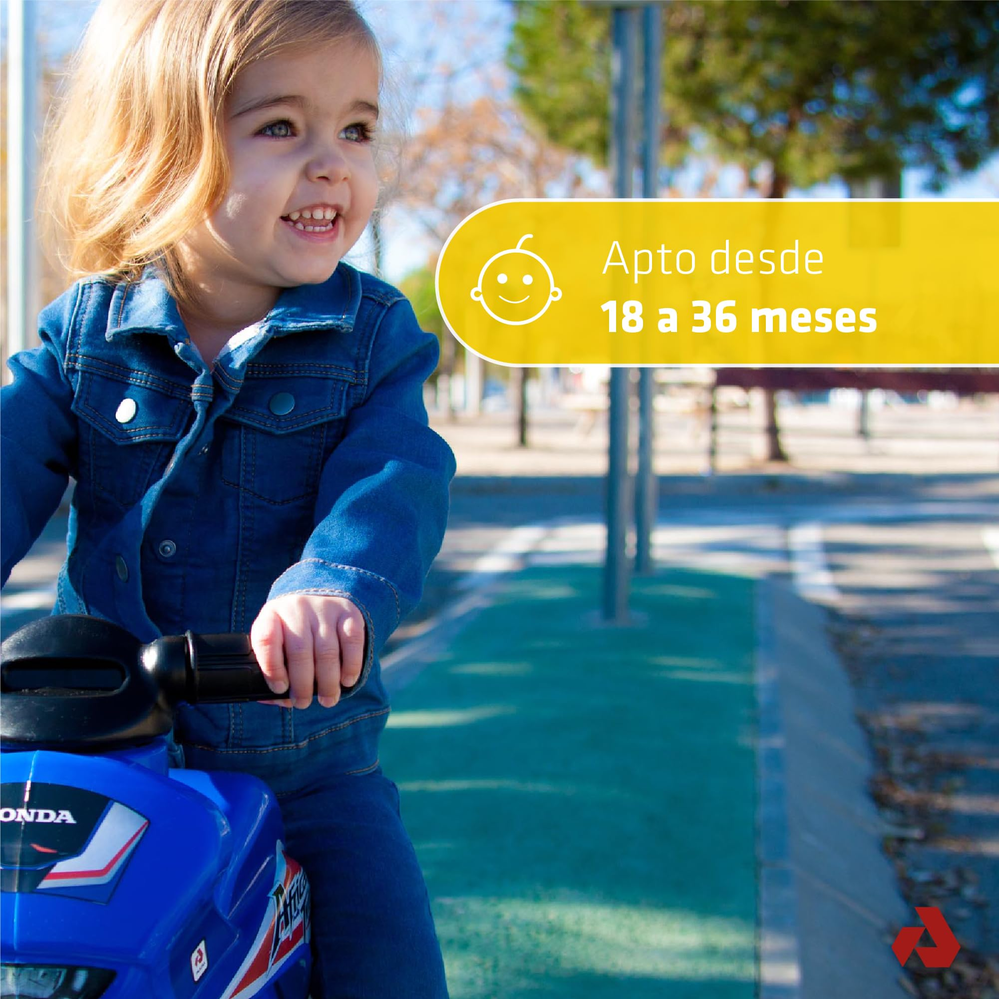 Un niño pequeño sonriente en una motocicleta de juguete azul. El niño lleva una chaqueta vaquera y está al aire libre. El fondo incluye una pancarta amarilla.