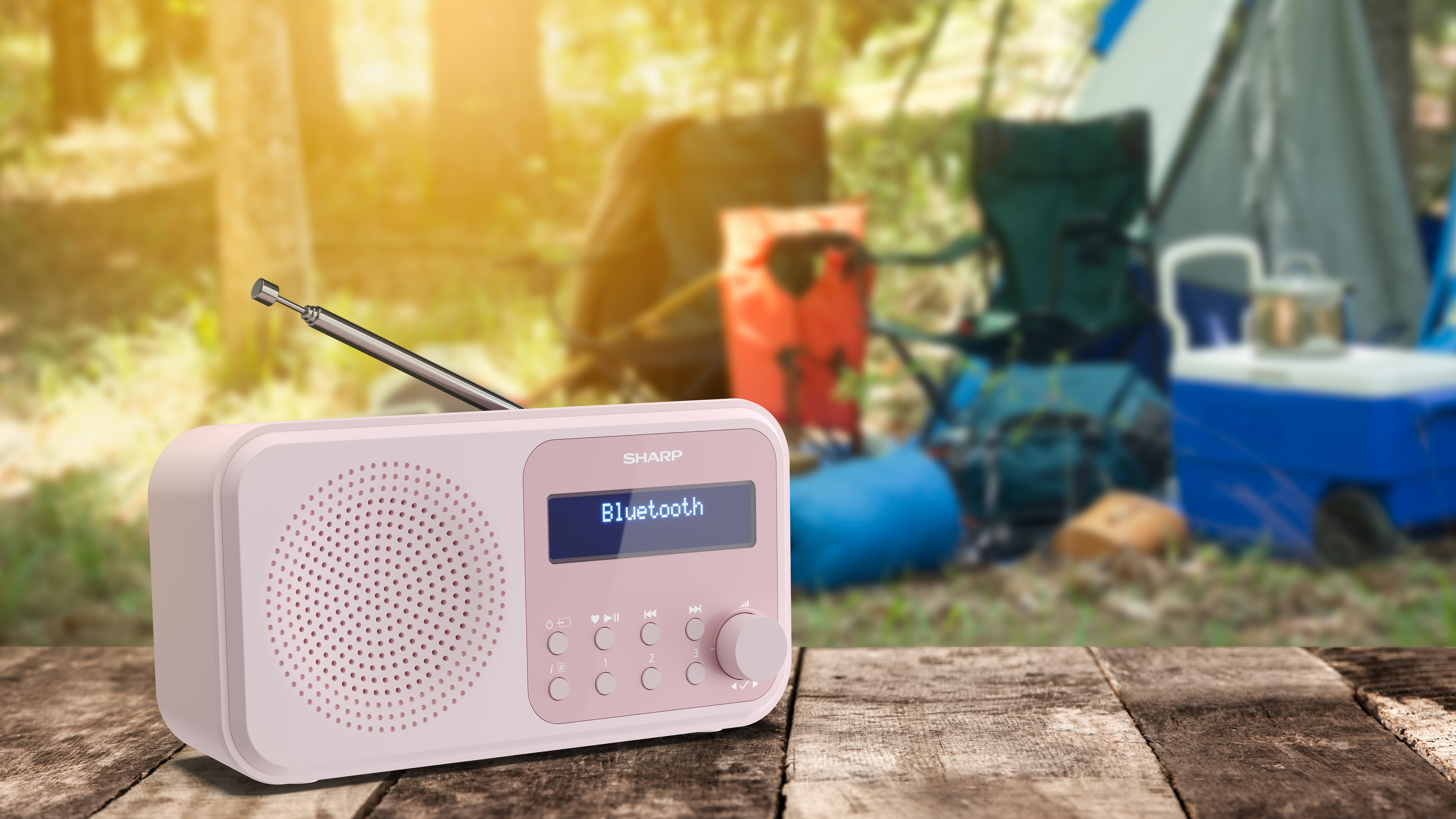 Une radio rose avec 'Bluetooth' à l'écran est posée sur du bois, équipement de camping flou en arrière-plan, tente, chaises.