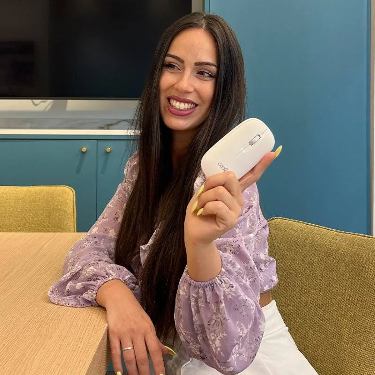 Una mujer sonríe, sosteniendo un ratón de computadora blanco. Lleva una camisa floral y pantalones blancos. El fondo tiene una pared azul y muebles.