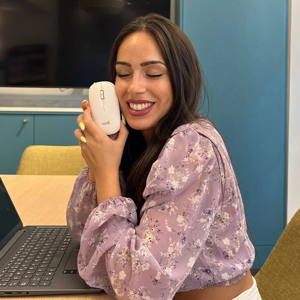 Mujer sonríe, sosteniendo un ratón blanco. Usa una blusa floral. Portátil en la mesa. Pared azul al fondo.