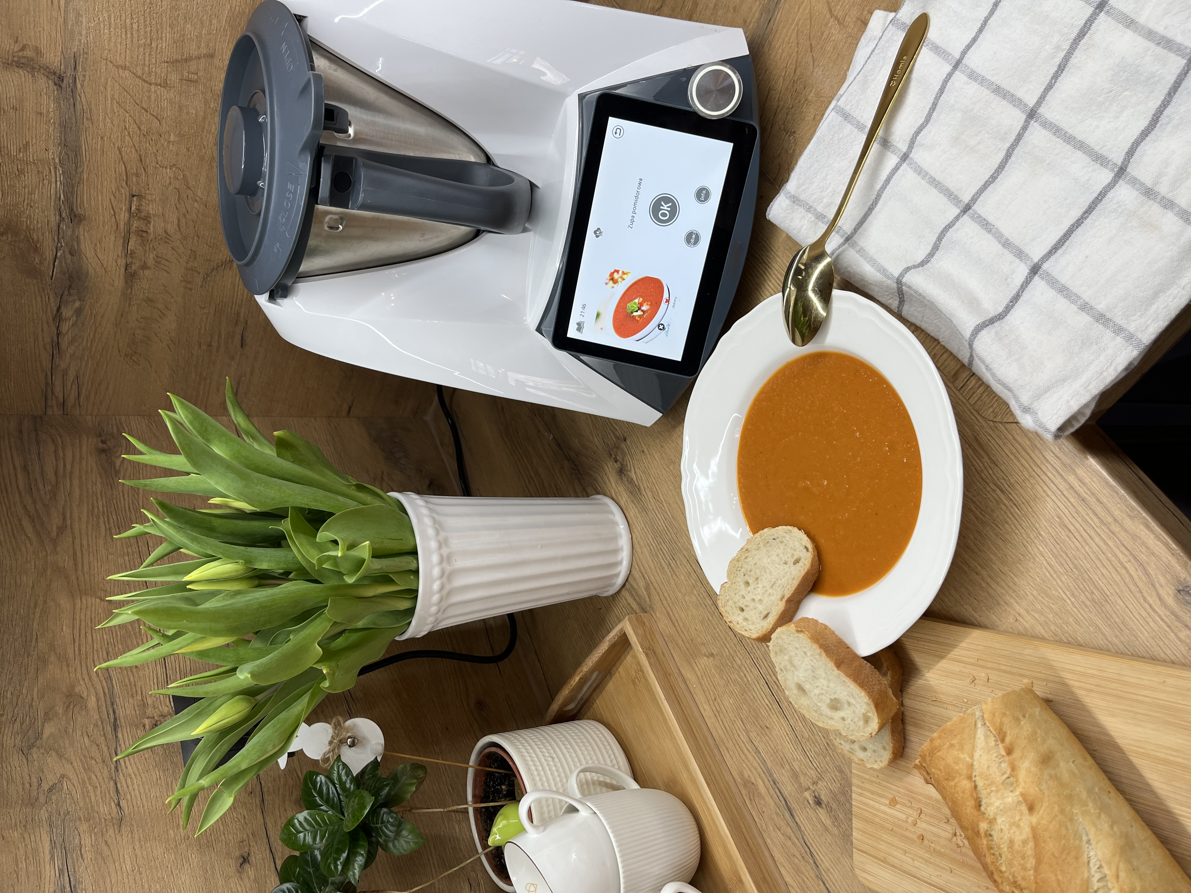 Scena kuchenna z robotem kuchennym, zupą, chlebem, kwiatami i łyżką na drewnianym stole.