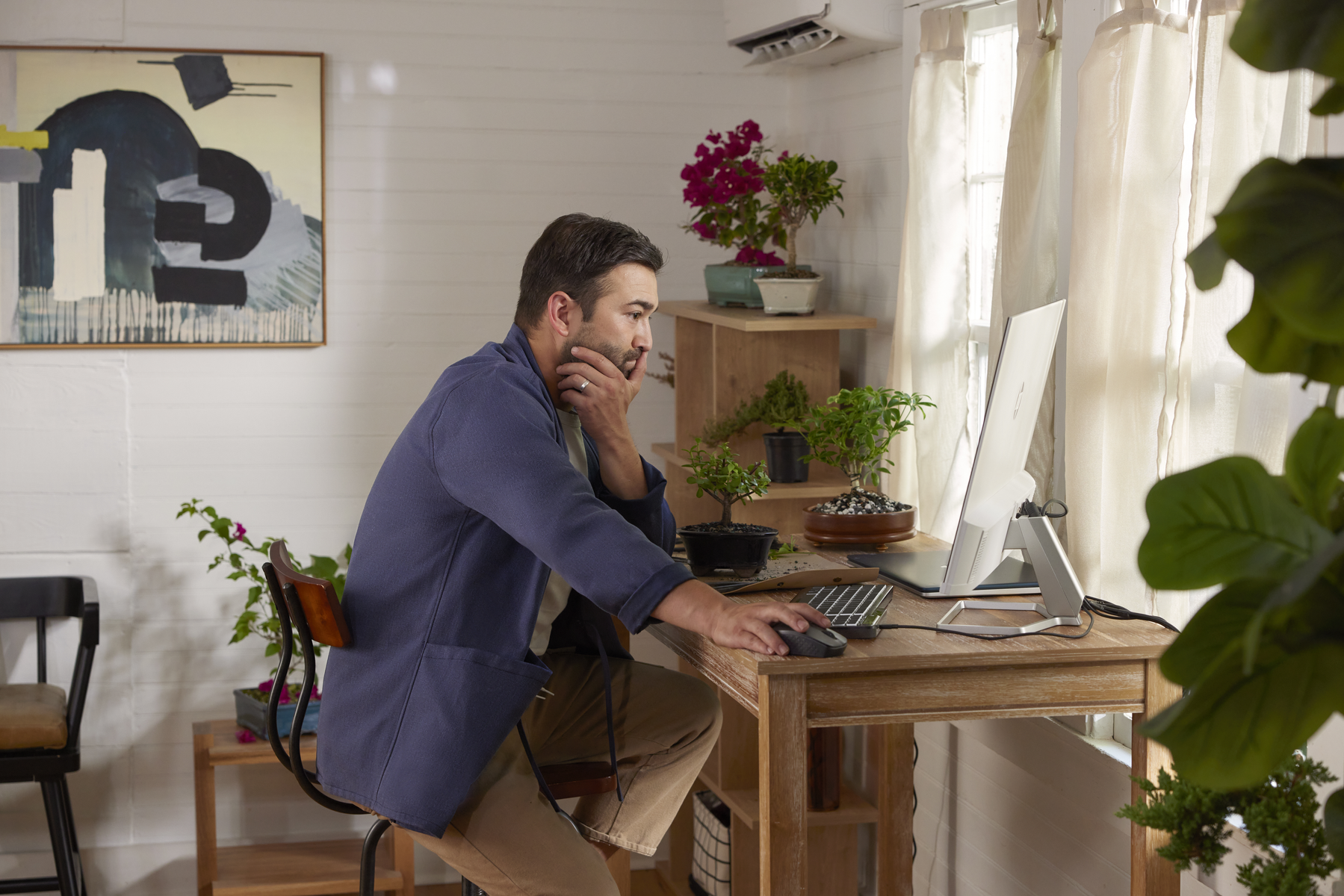 Un hombre con una chaqueta azul trabaja en una computadora. Hay plantas, una pintura y una ventana detrás de él.