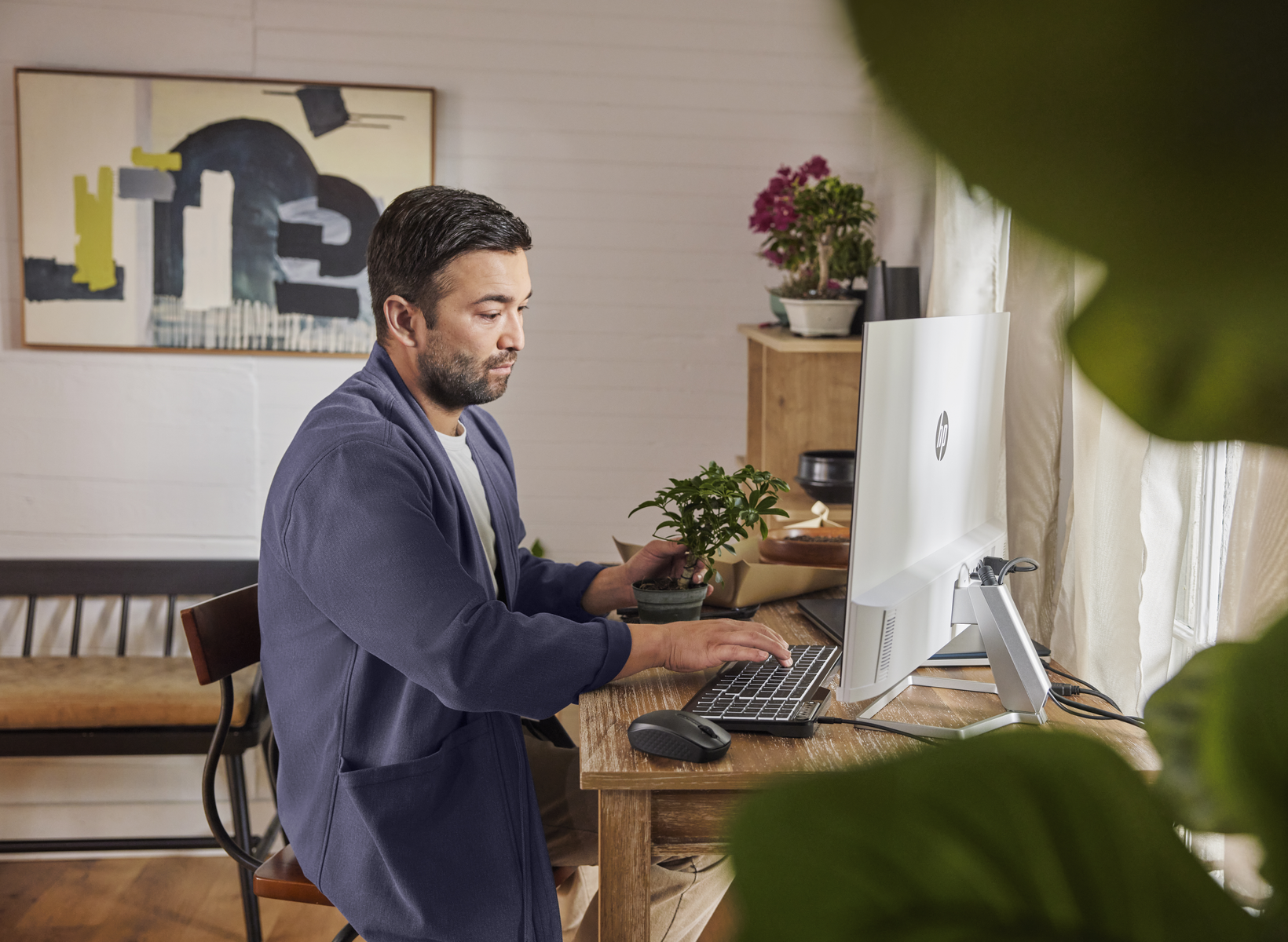 Hombre con bata azul trabaja en computadora con planta. Escritorio con ratón y teclado. Fondo con arte y flores.
