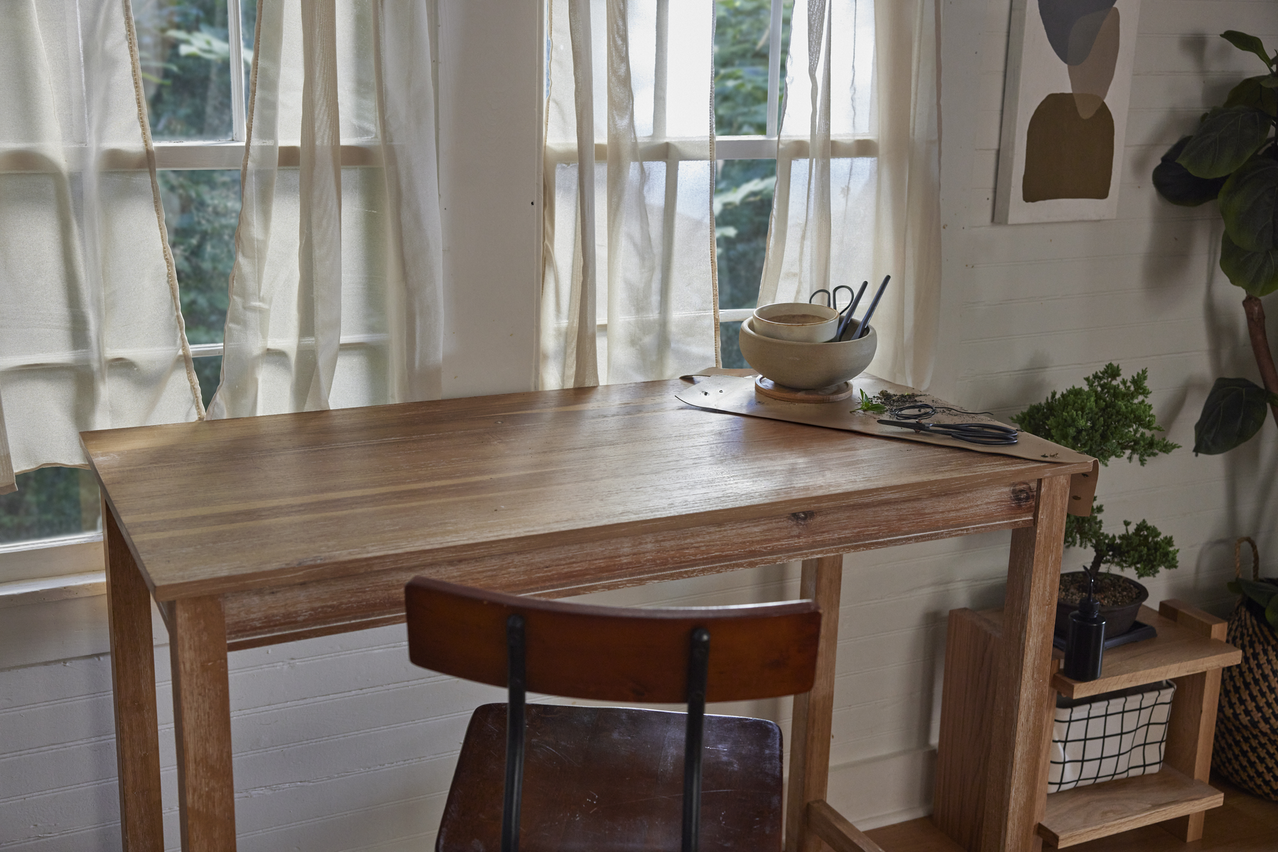 Escritorio de madera con cuenco, tijeras y vegetación. Una silla al frente. Ventana con cortinas transparentes al fondo.