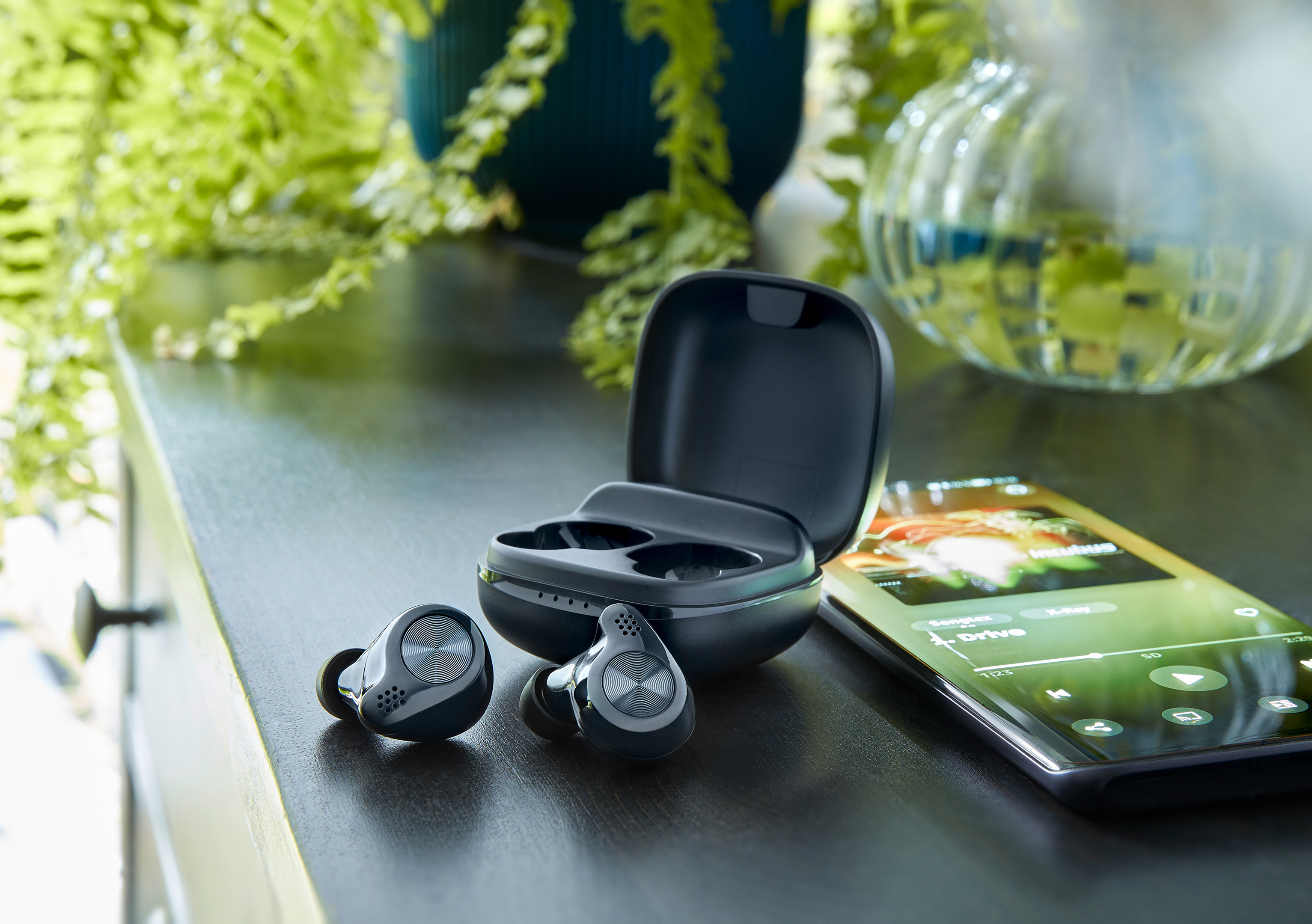 Auricolari, custodia e smartphone su una superficie in legno scuro, con piante sullo sfondo.