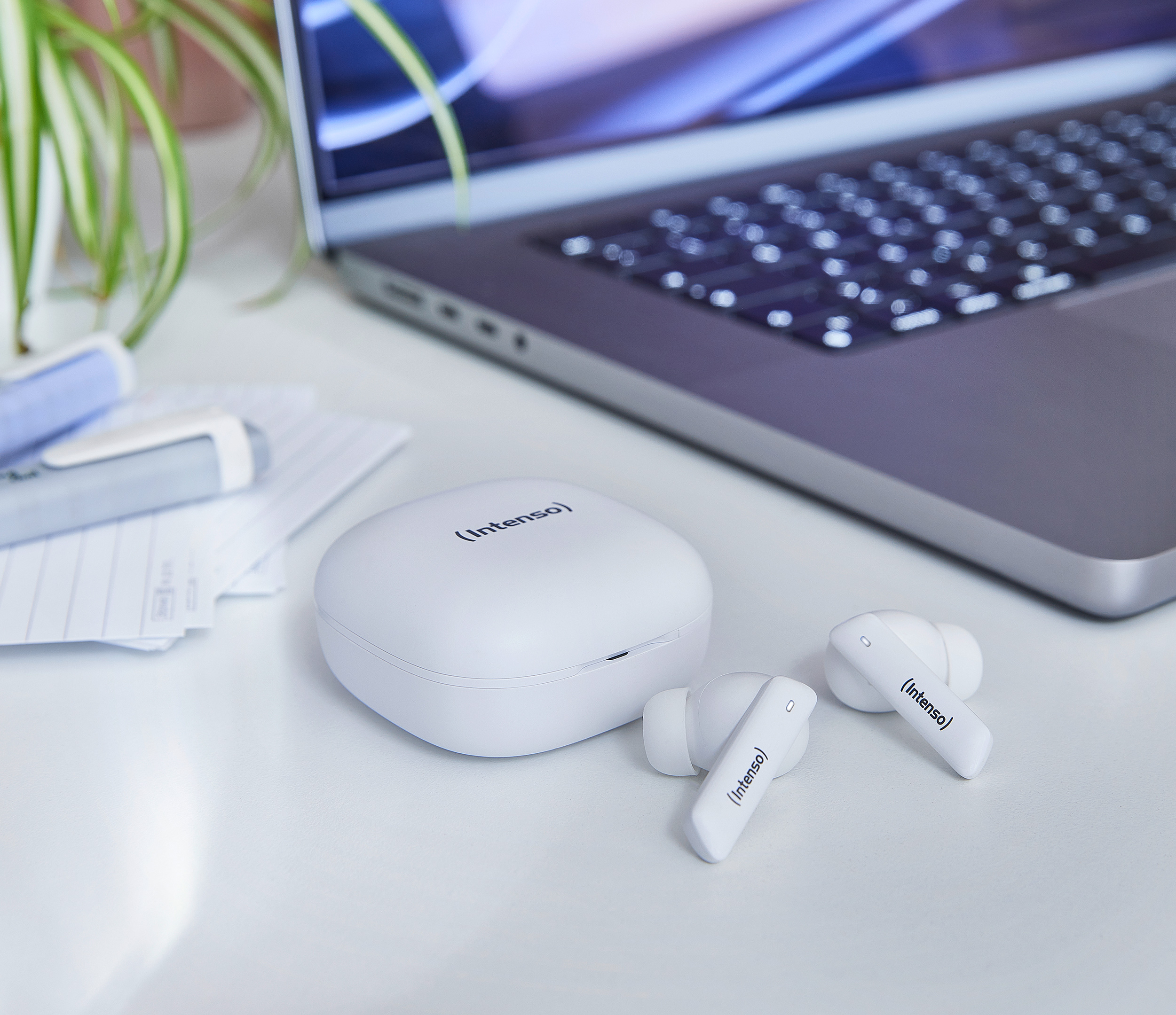Écouteurs Intenso blancs, étui de chargement et ordinateur portable sur un bureau blanc. Des papiers et une plante sont également visibles.