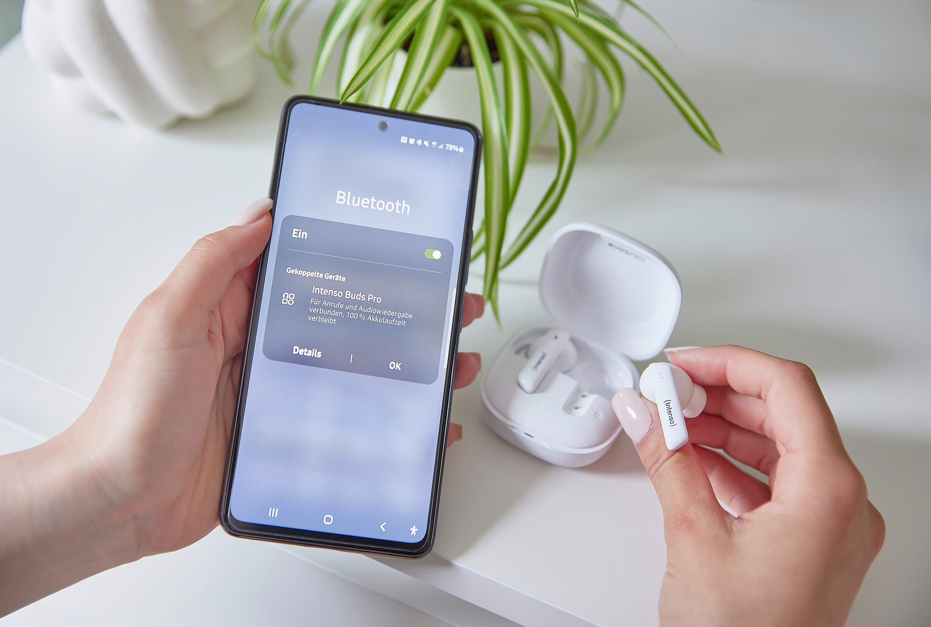 Un téléphone affiche les paramètres Bluetooth, connecté aux Intenso Buds Pro. Une main tient un écouteur blanc, l'autre le téléphone.