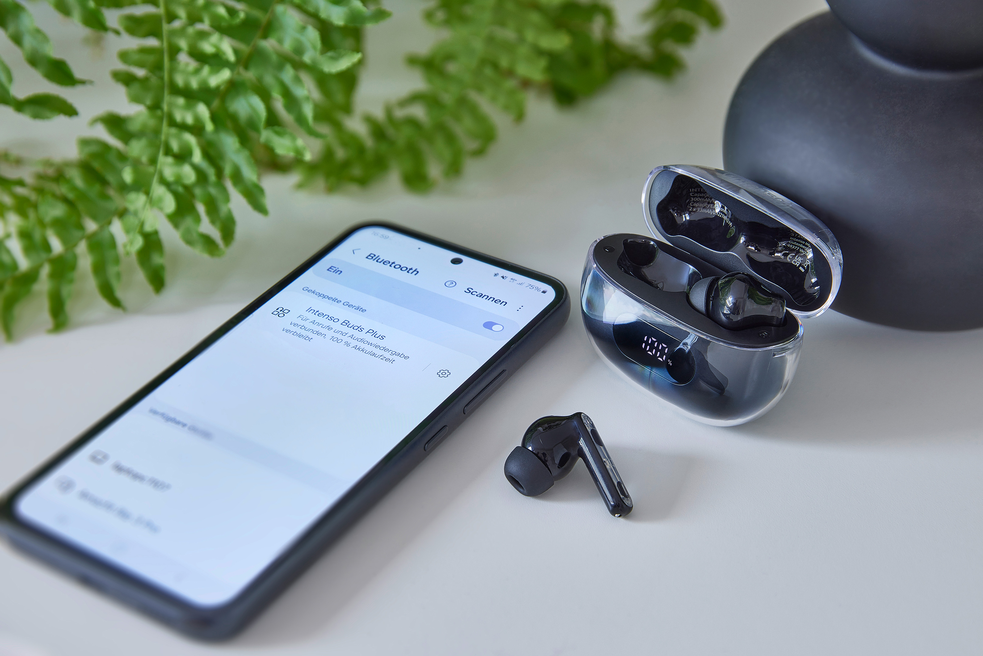 Smartfon z ustawieniami Bluetooth, bezprzewodowe słuchawki i etui ładujące na białej powierzchni z zielonymi liśćmi.