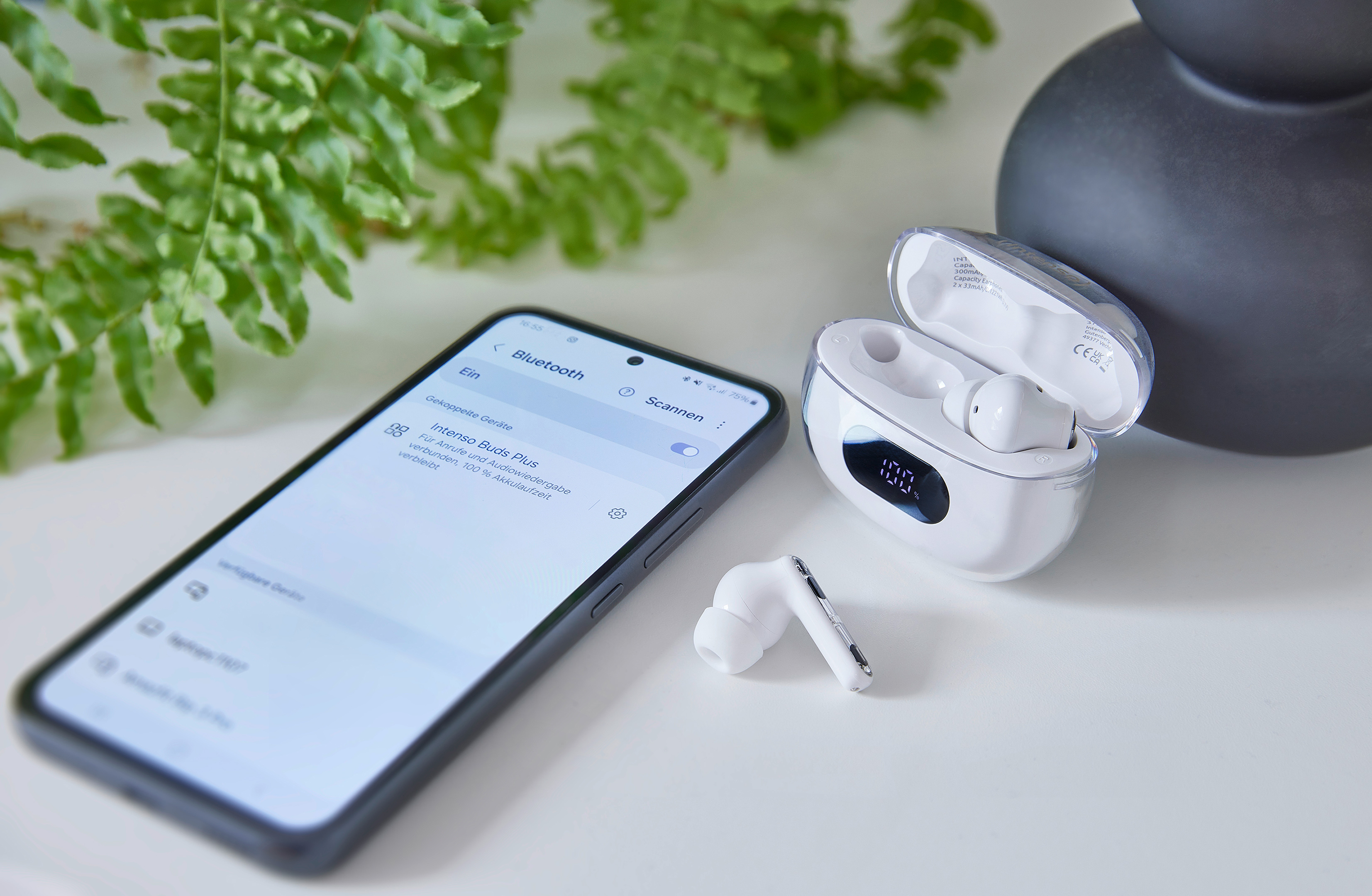 Smartphone mit Bluetooth-Einstellungen, Ohrhörer und Ladecase auf weißer Oberfläche mit grüner Pflanze im Hintergrund.