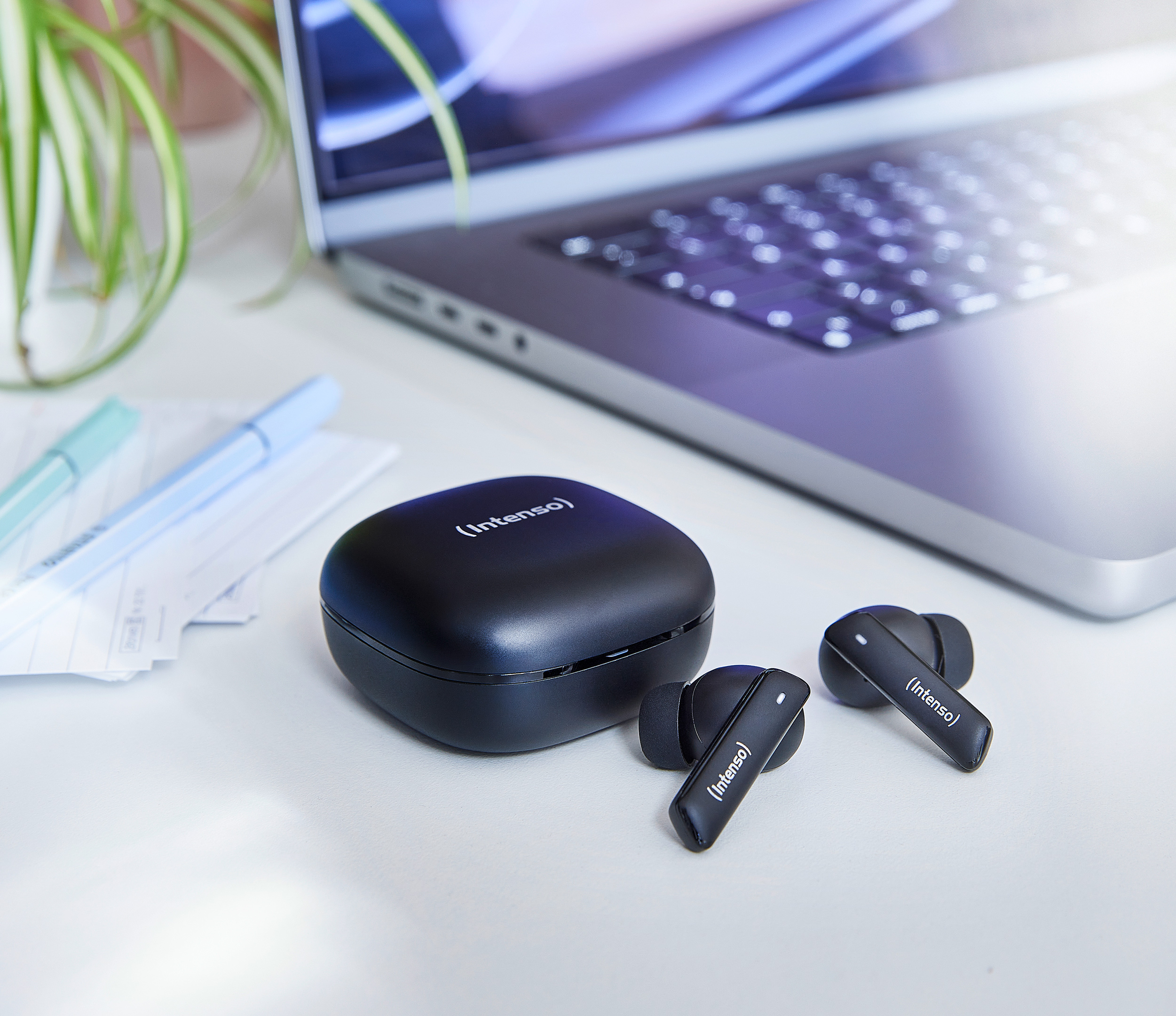 Una escena de escritorio con una computadora portátil, auriculares Intenso, estuche, bolígrafos y una planta. Los auriculares son negros.