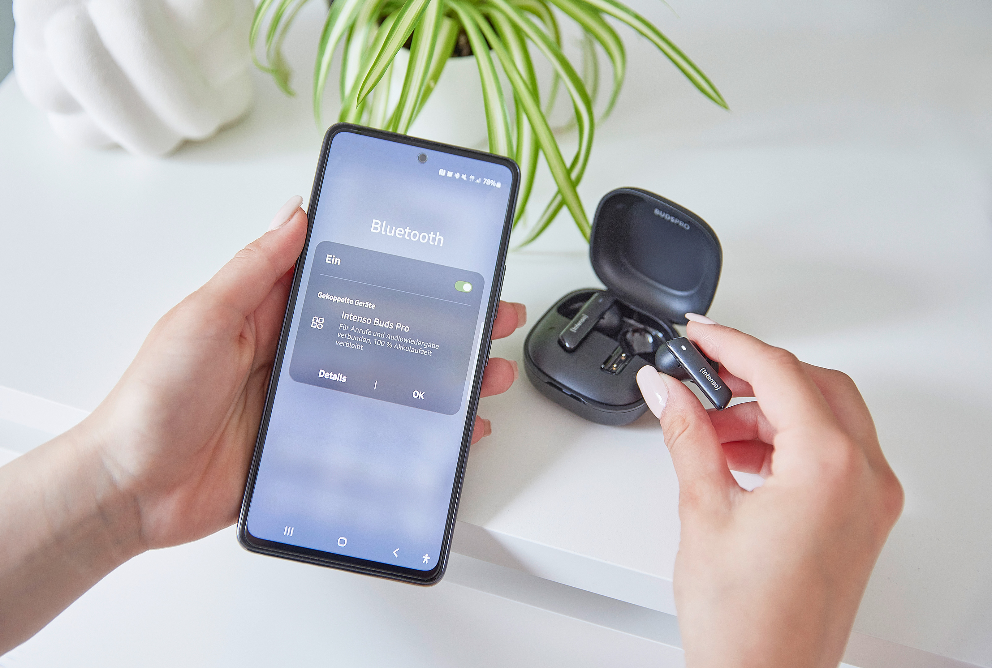 Una persona sostiene un teléfono con ajustes de Bluetooth. Auriculares y su estuche sobre una superficie blanca.
