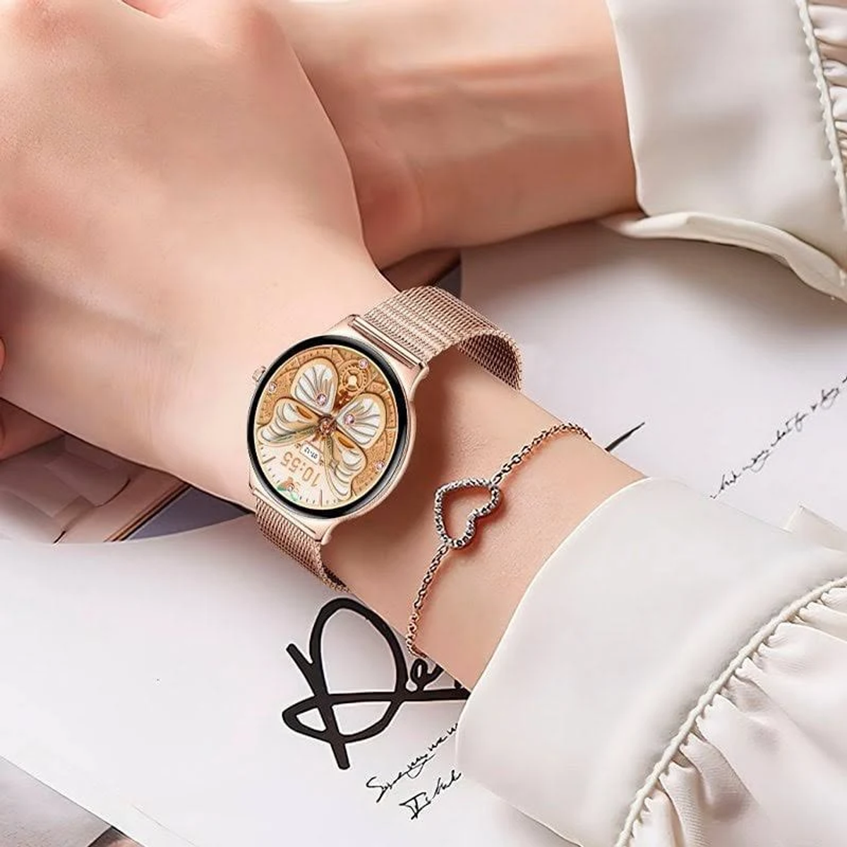 Il polso di una donna con un orologio in oro rosa e un braccialetto a cuore. Il quadrante dell'orologio ha un design a farfalla. La donna indossa una camicia bianca.