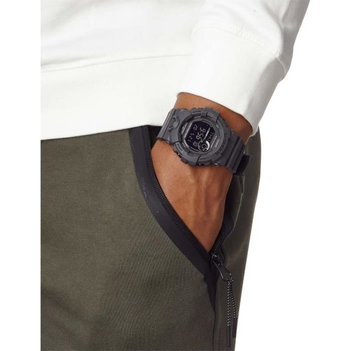 Una persona indossa un orologio grigio al polso sinistro. Indossa una camicia bianca e pantaloni verdi con tasca.