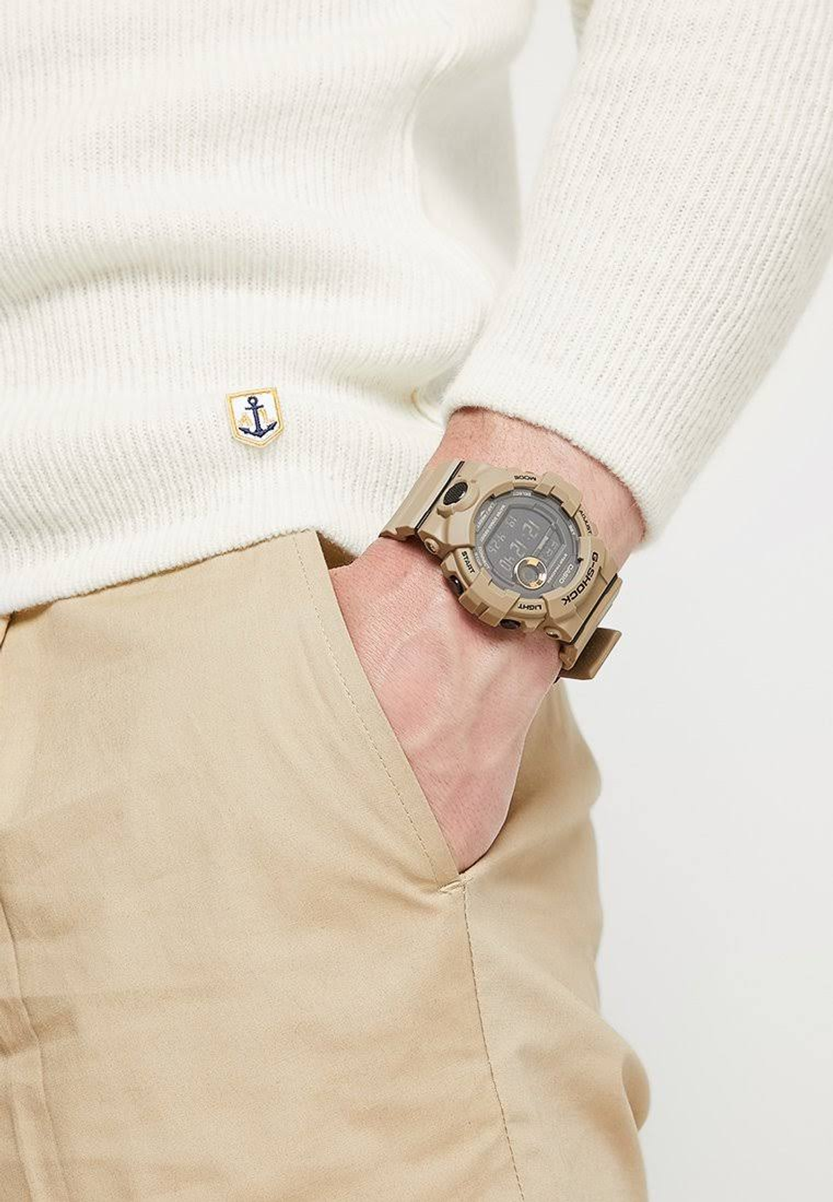 Una persona lleva un suéter crema con un parche de ancla, pantalones caqui y un reloj G-Shock color canela, con la mano en el bolsillo.