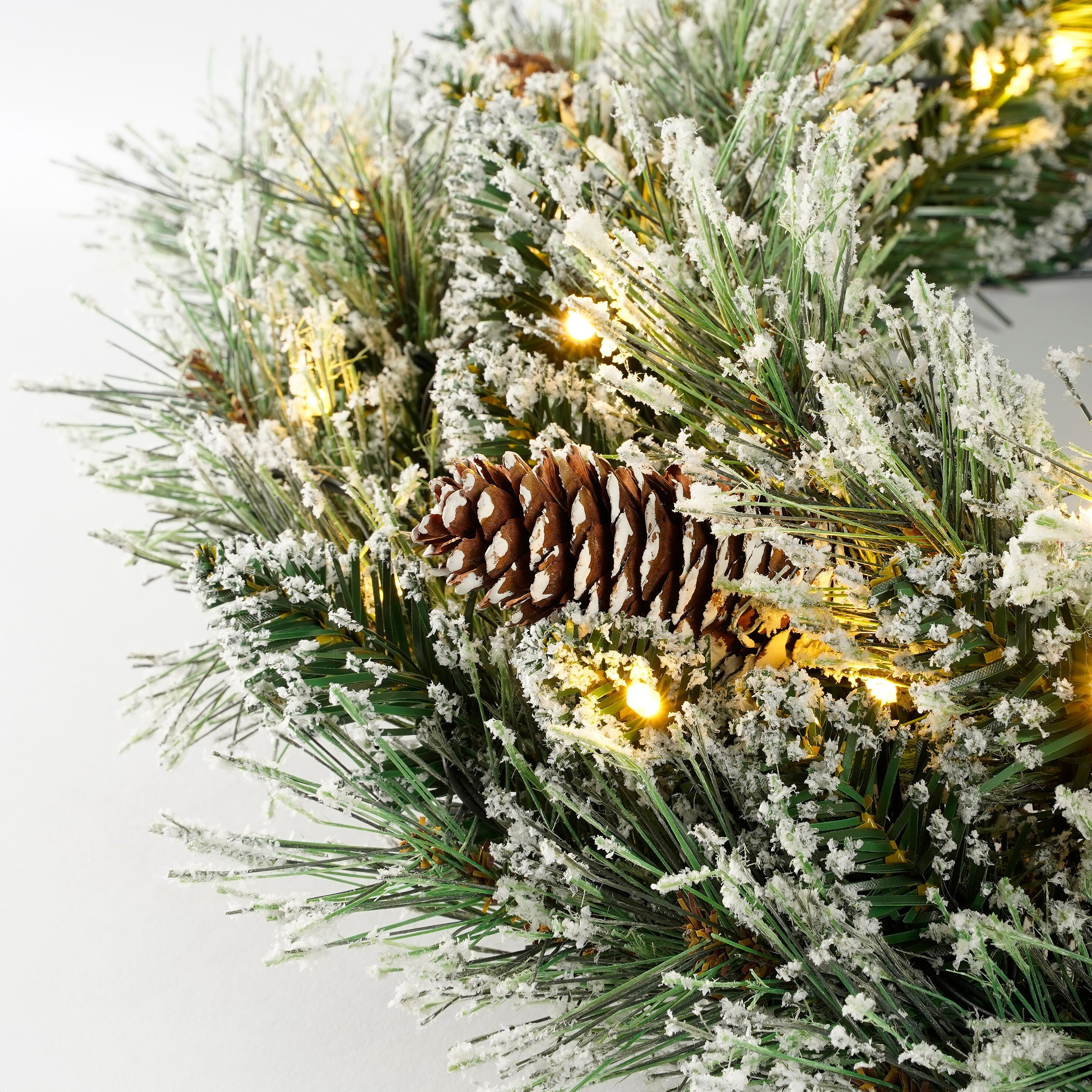 Gros plan d'une couronne de Noël avec des aiguilles de pin, de la neige et une pomme de pin. Lumières visibles.
