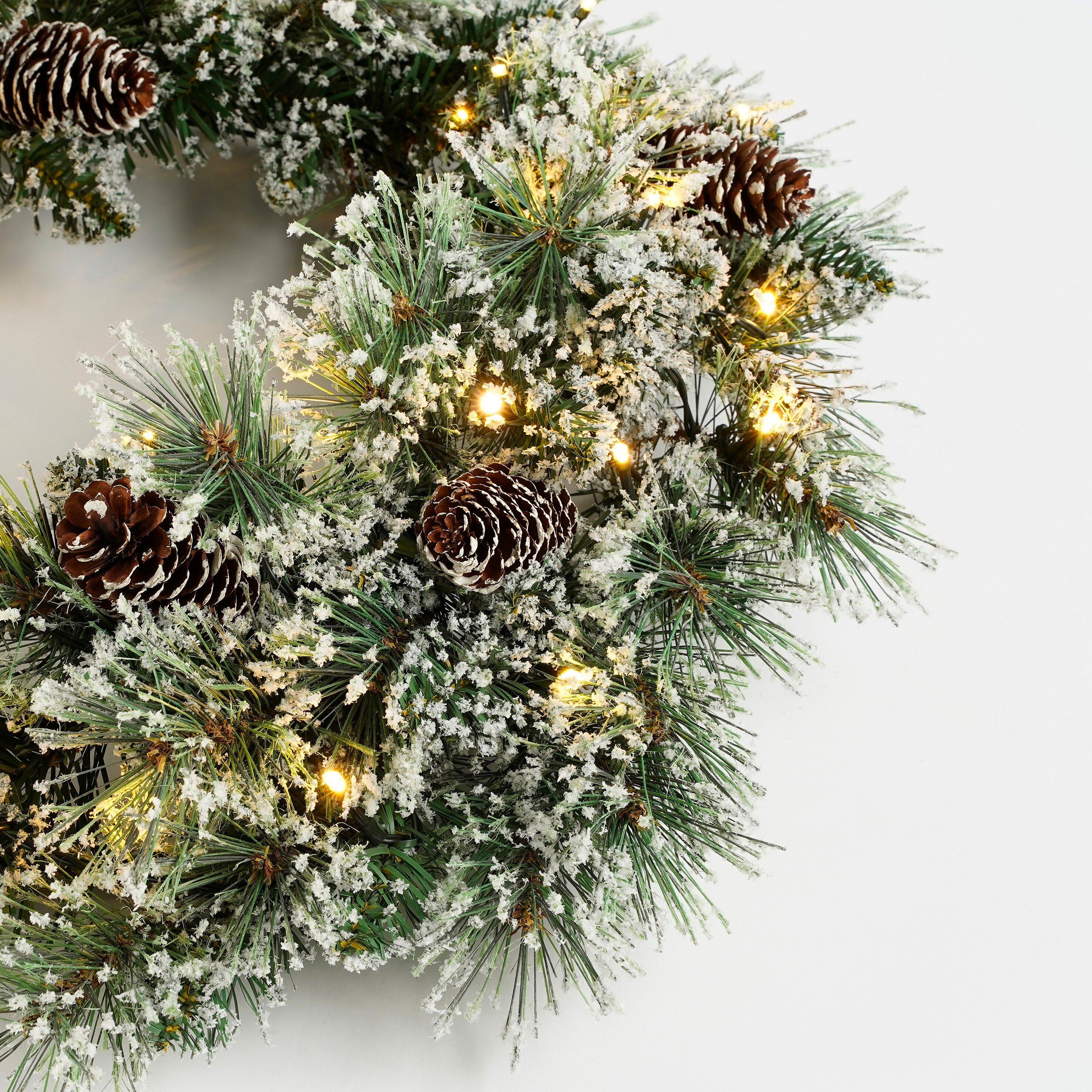 Une couronne de Noël avec des aiguilles de pin, de la neige et des pommes de pin, éclairée par de petites lumières.