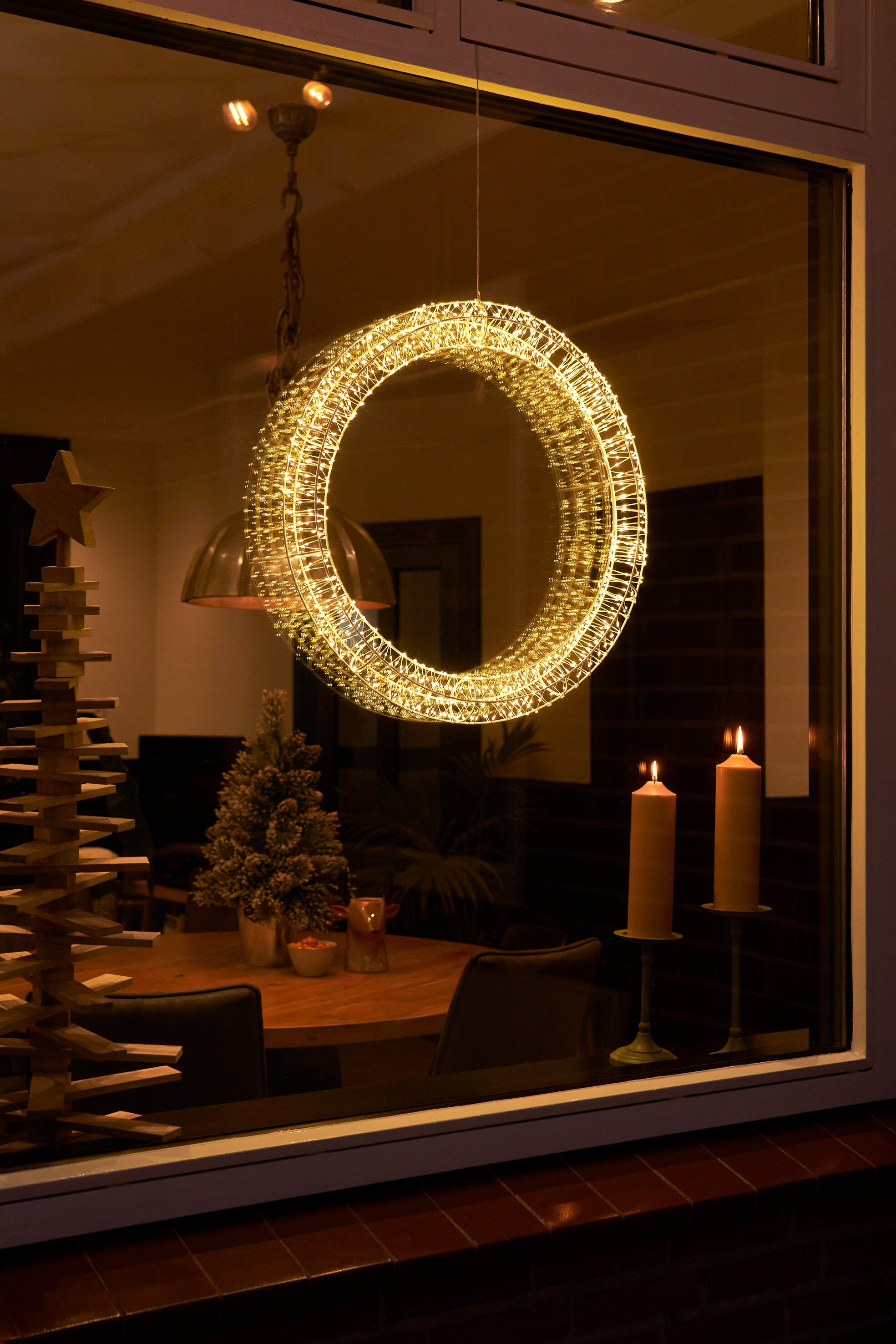 Une couronne de Noël éclairée est suspendue dans une fenêtre. À l'intérieur, bougies et arbre en bois.