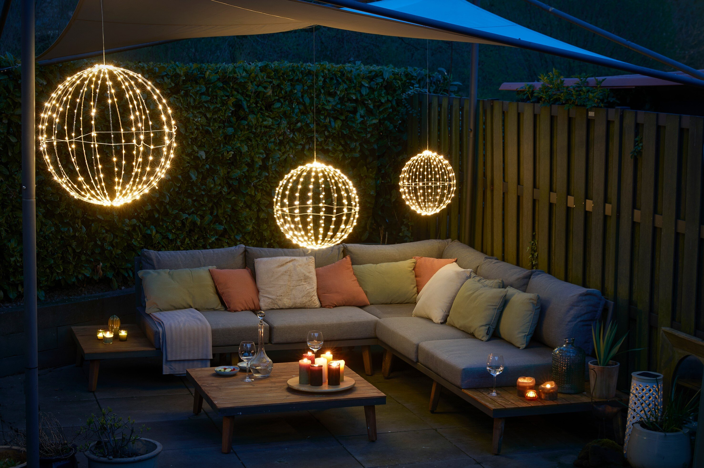 Patio extérieur avec canapé gris, éclairé par des sphères lumineuses suspendues. Bougies et coussins.