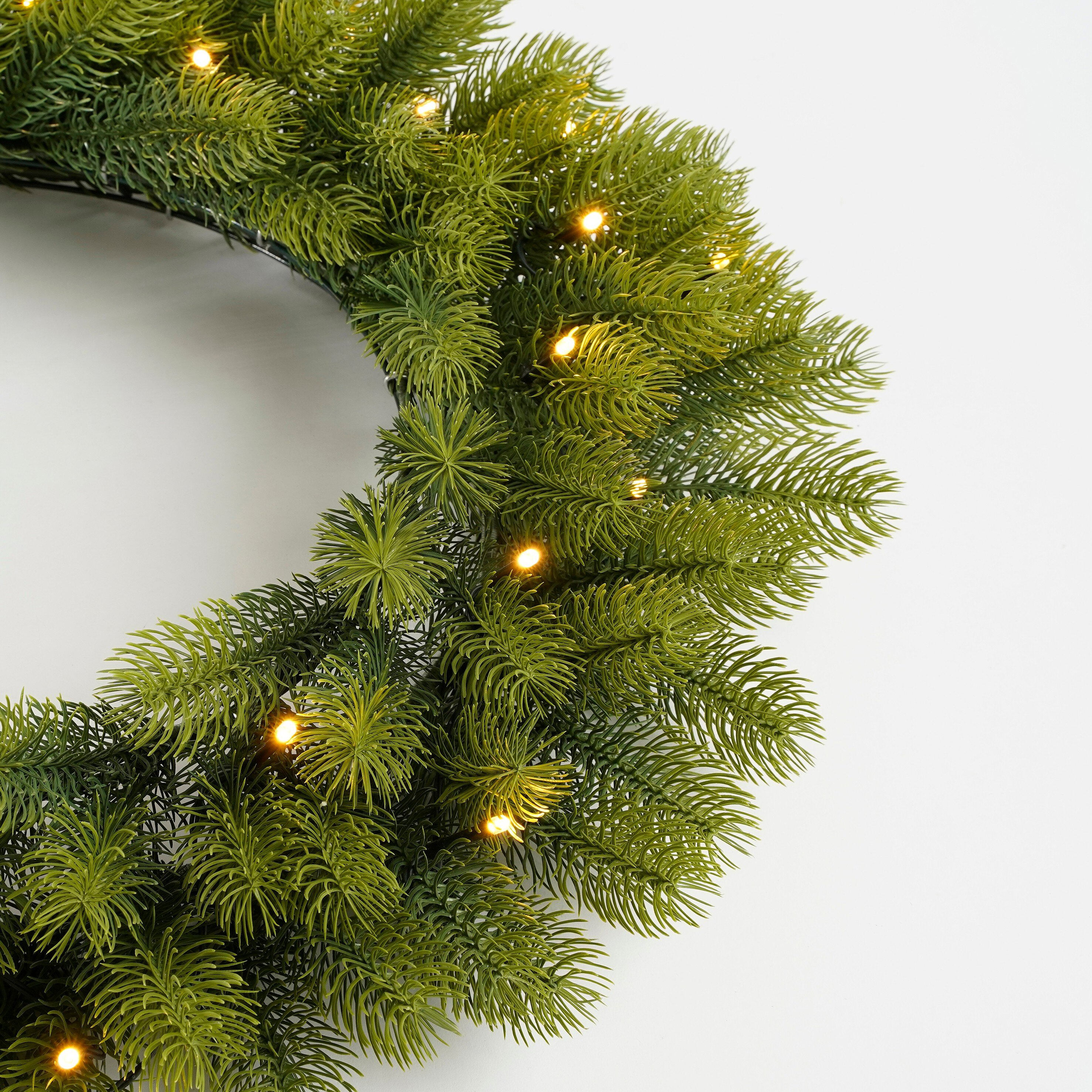 Une couronne de Noël verte avec de petites lumières, sur fond blanc. La couronne est éclairée.
