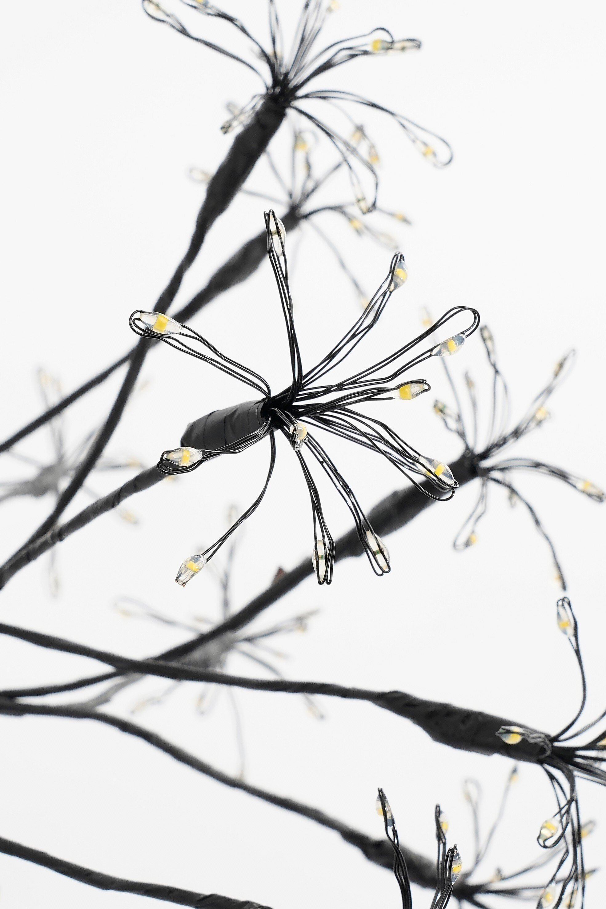 Arbre décoratif noir avec de petites lumières sur fond blanc. Les lumières sont jaunes. Les branches sont en forme de fil.