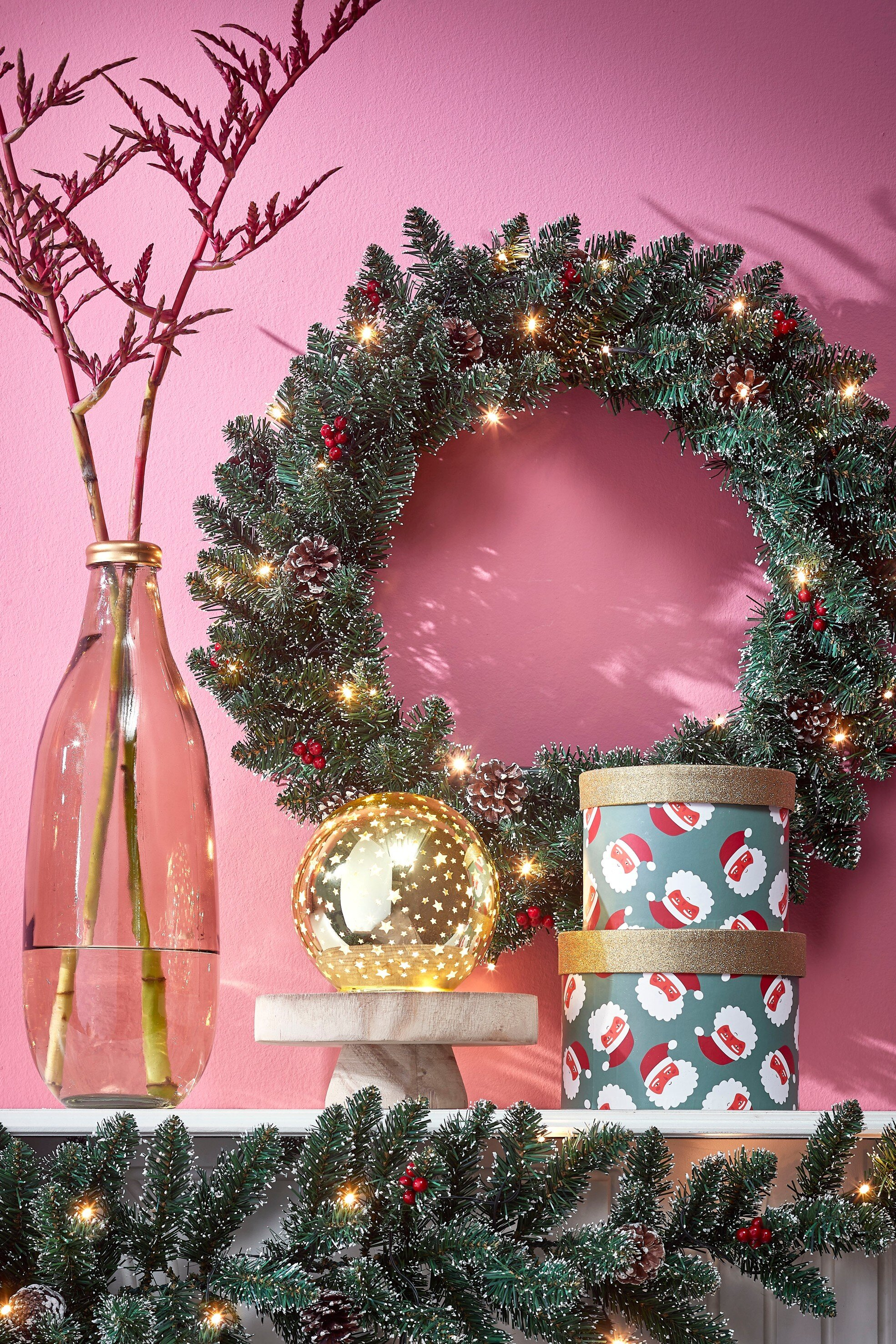 Décorations de Noël : couronne, boîtes Père Noël, globe étoilé, vase avec branches et guirlande sur fond rose.