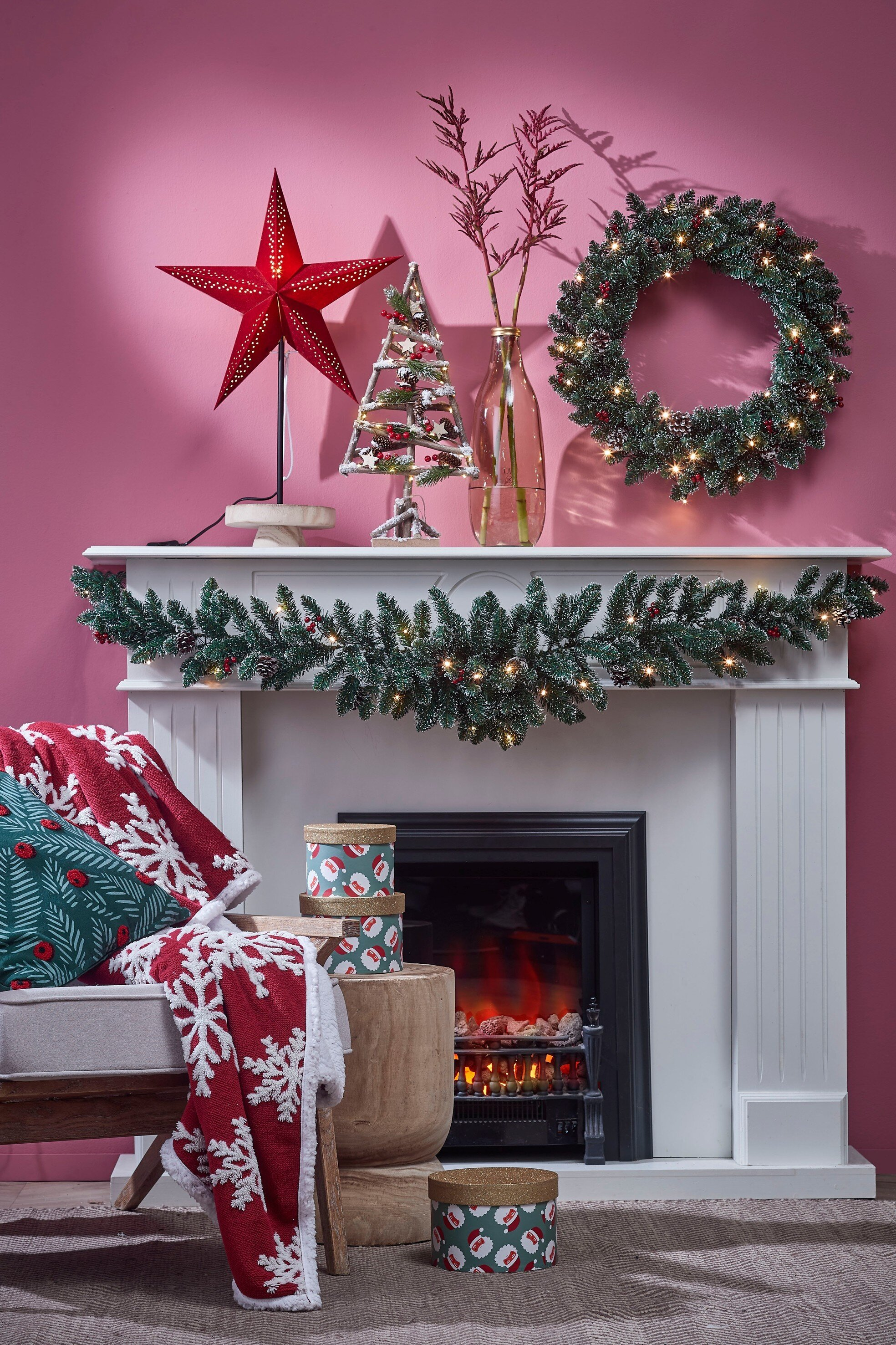 Une cheminée décorée avec une étoile de Noël, une couronne, une guirlande et des cadeaux. Une couverture et un coussin sont sur une chaise.