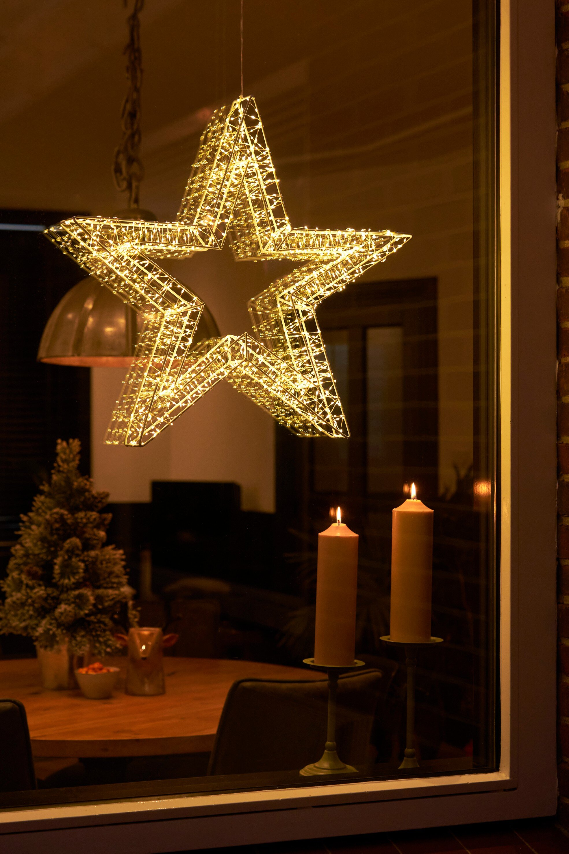 Une étoile lumineuse décorative est suspendue à une fenêtre. Deux bougies allumées. Petit sapin et table.