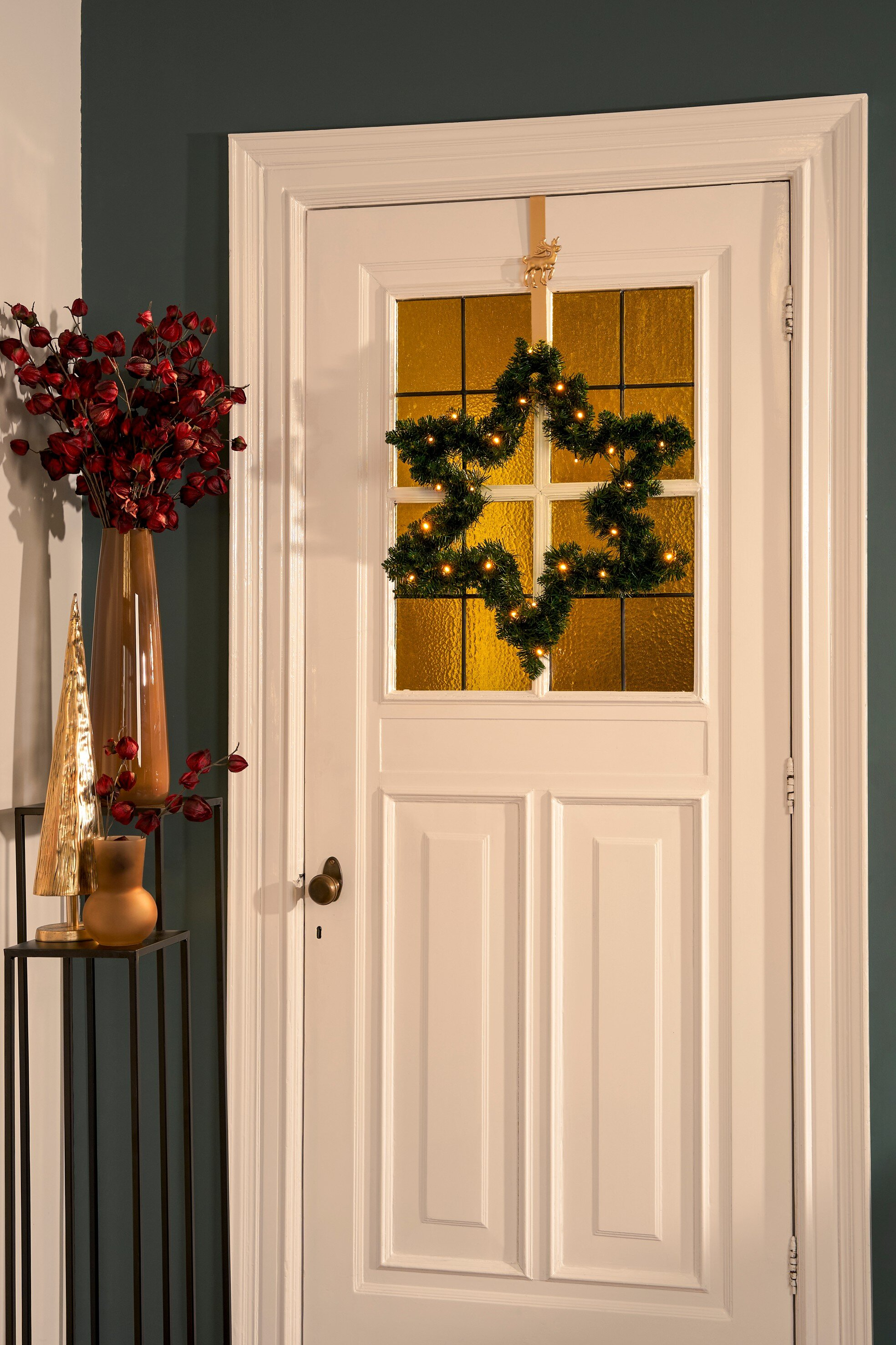 Une porte blanche avec une couronne de Noël en forme d'étoile. Un vase avec des fleurs rouges à gauche.