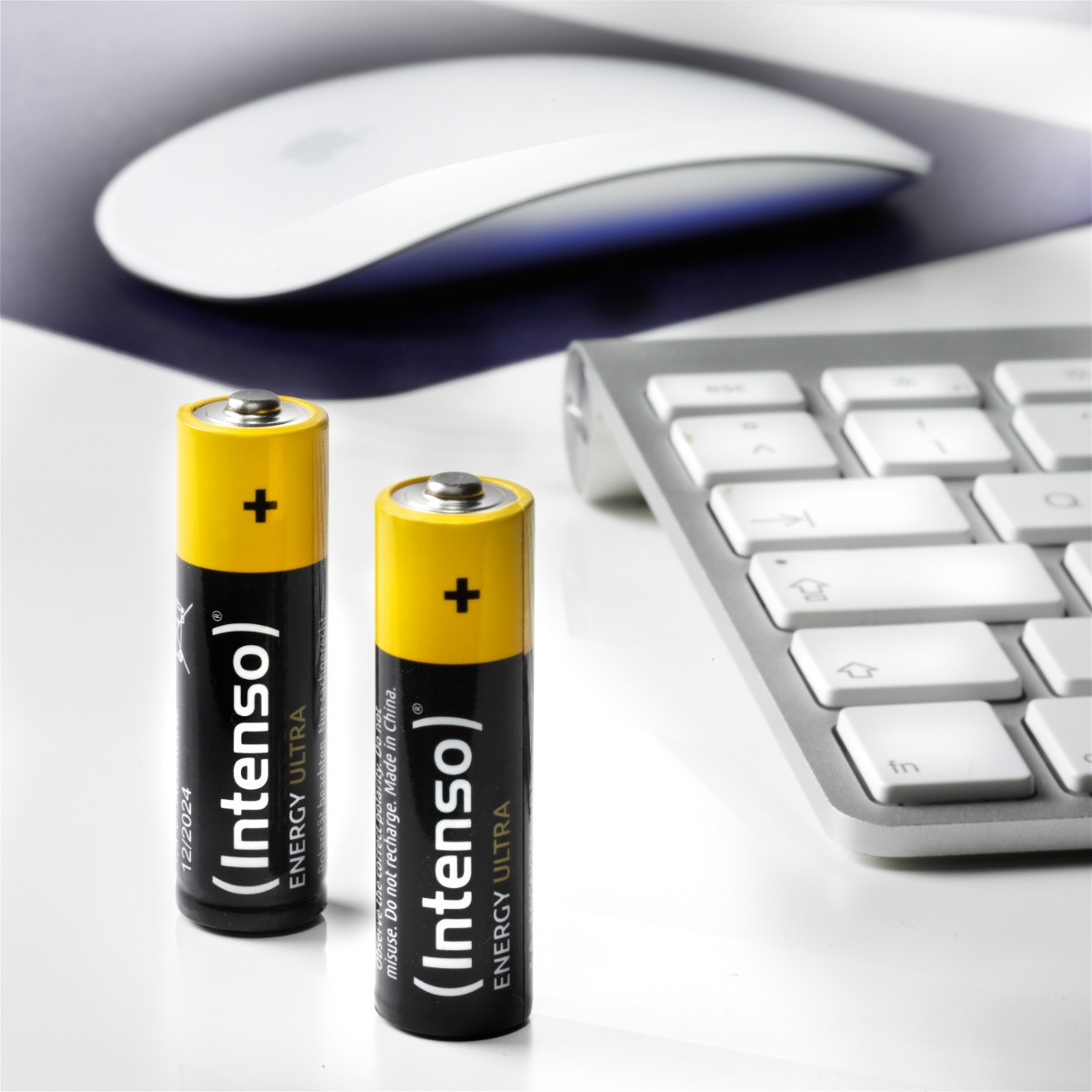 Zwei Batterien mit gelben Oberteilen und schwarzen Etiketten, eine weiße Maus und eine Tastatur auf weißer Oberfläche.