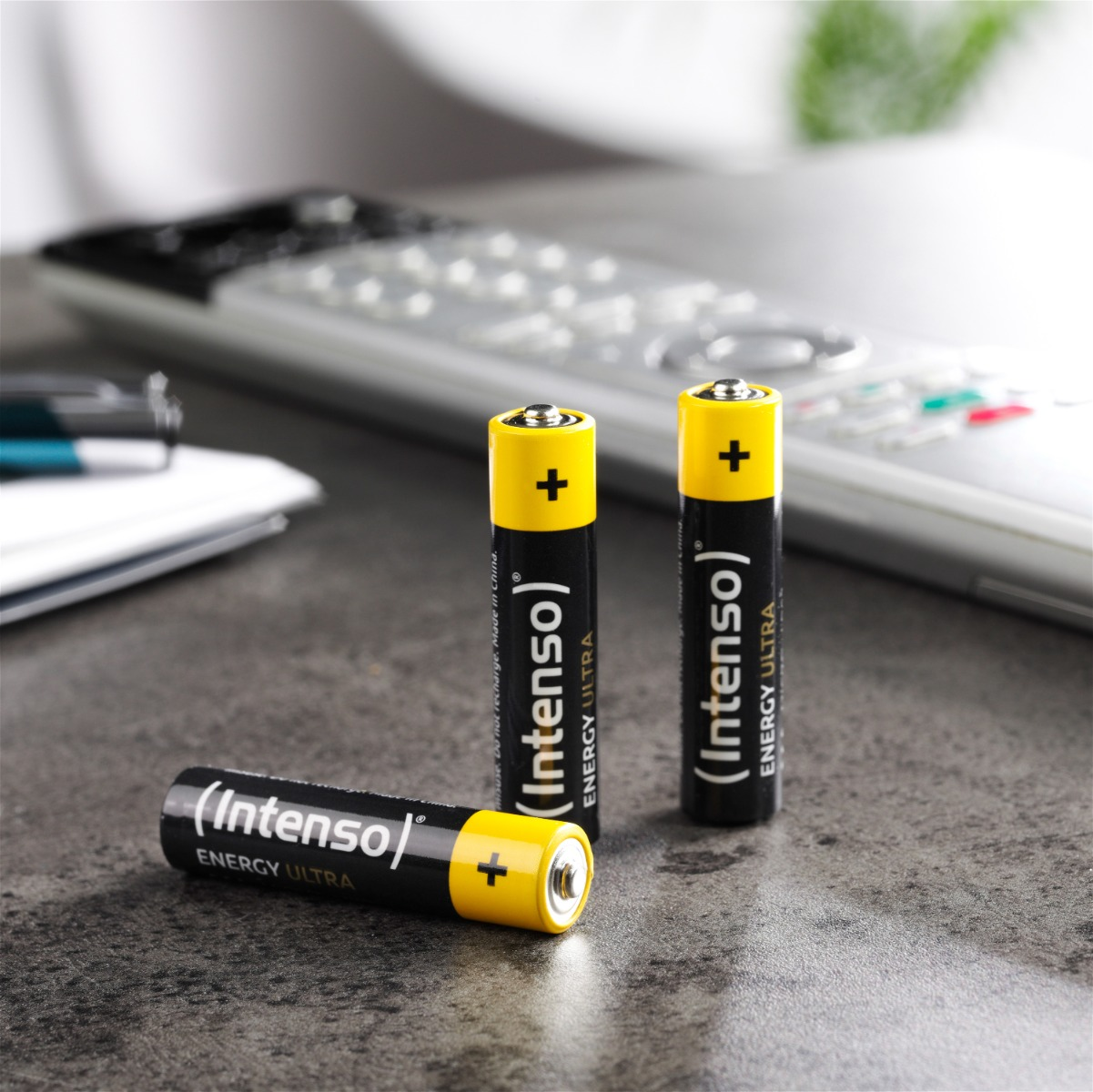 Tre batterie AA, parte superiore gialla con segno più, corpo nero con testo bianco, su una superficie grigia, con un telecomando sullo sfondo.