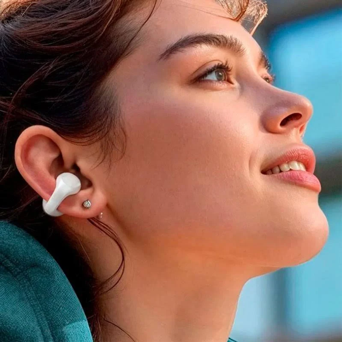 Una mujer mira hacia arriba, con un auricular blanco. Tiene cabello castaño, piel clara y un pequeño pendiente. Se ve un fondo azul.