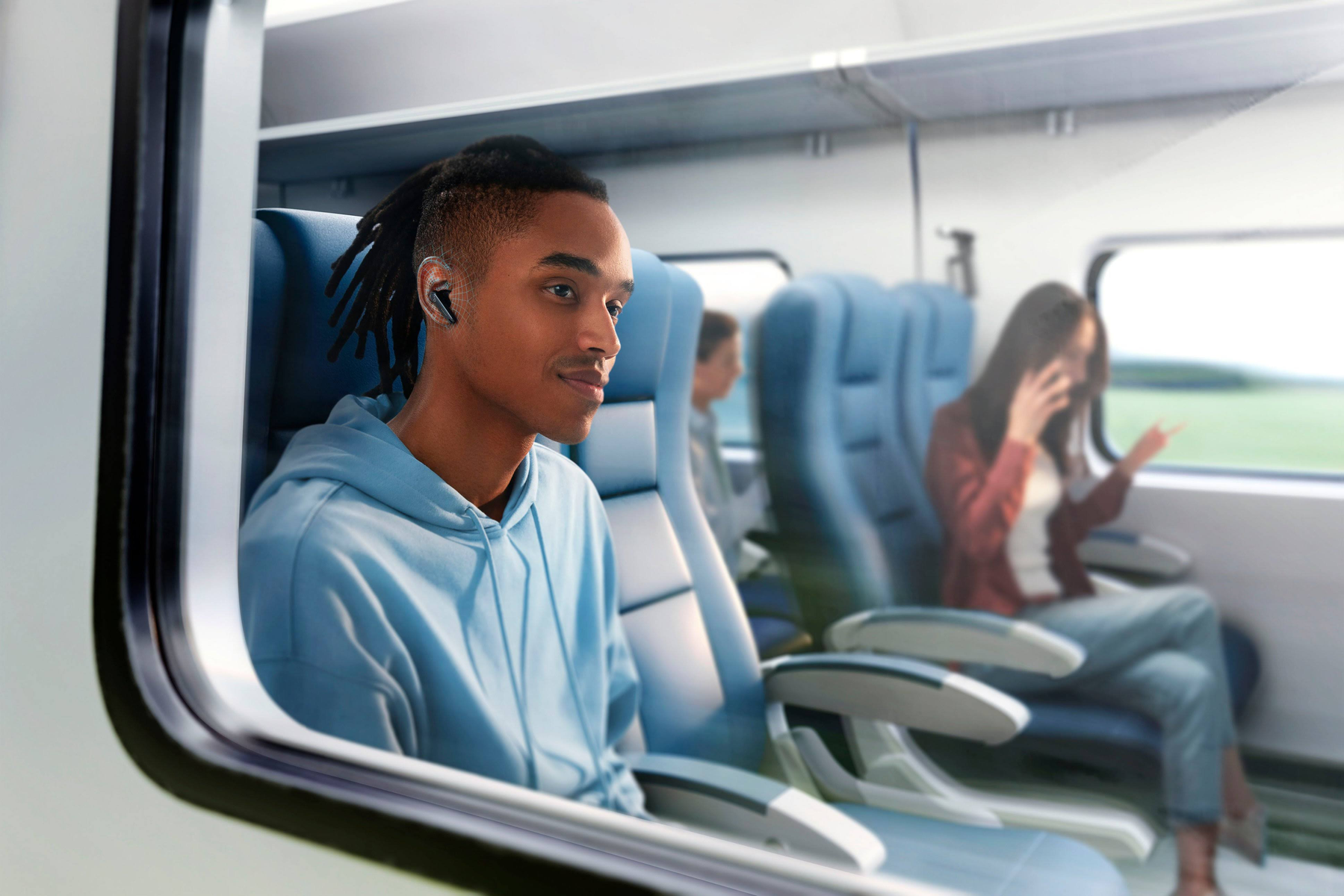 Un uomo con una felpa blu è in treno, ascoltando musica. Un'altra donna è al telefono. Fuori, campi verdi.